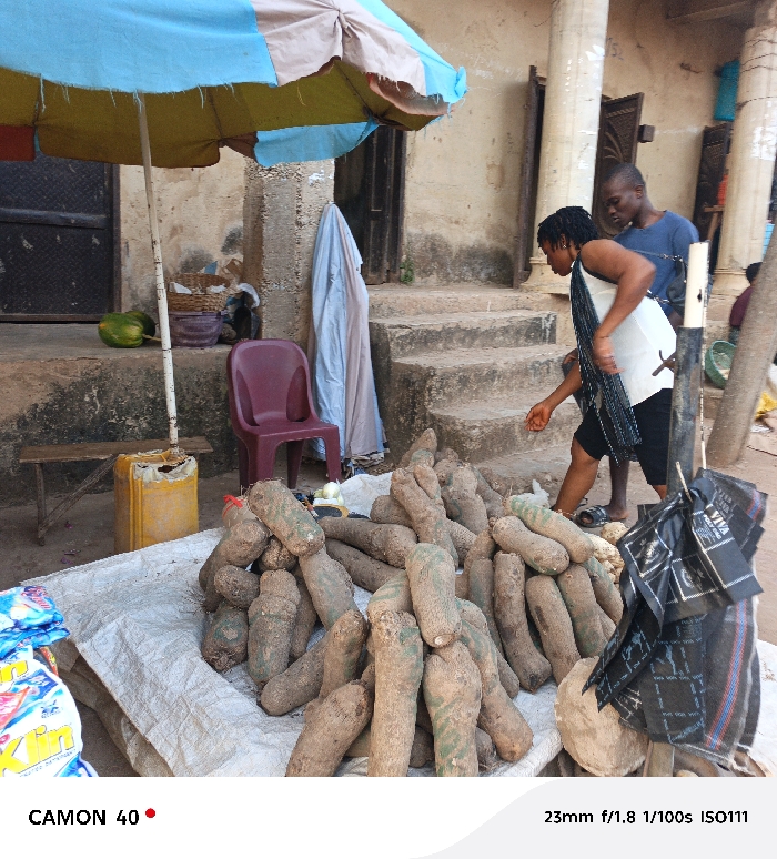 A vendor's stall displaying a large pile of yams covered by a white sheet. A person dressed in a white tank top and shorts is about to pick one up with a man standing nearby. Behind the stall, there's a weathered building with a small set of steps leading to a doorway. A multi-colored umbrella provides shade to the yams and the stall. A maroon plastic chair and a yellow canister are near the stall.