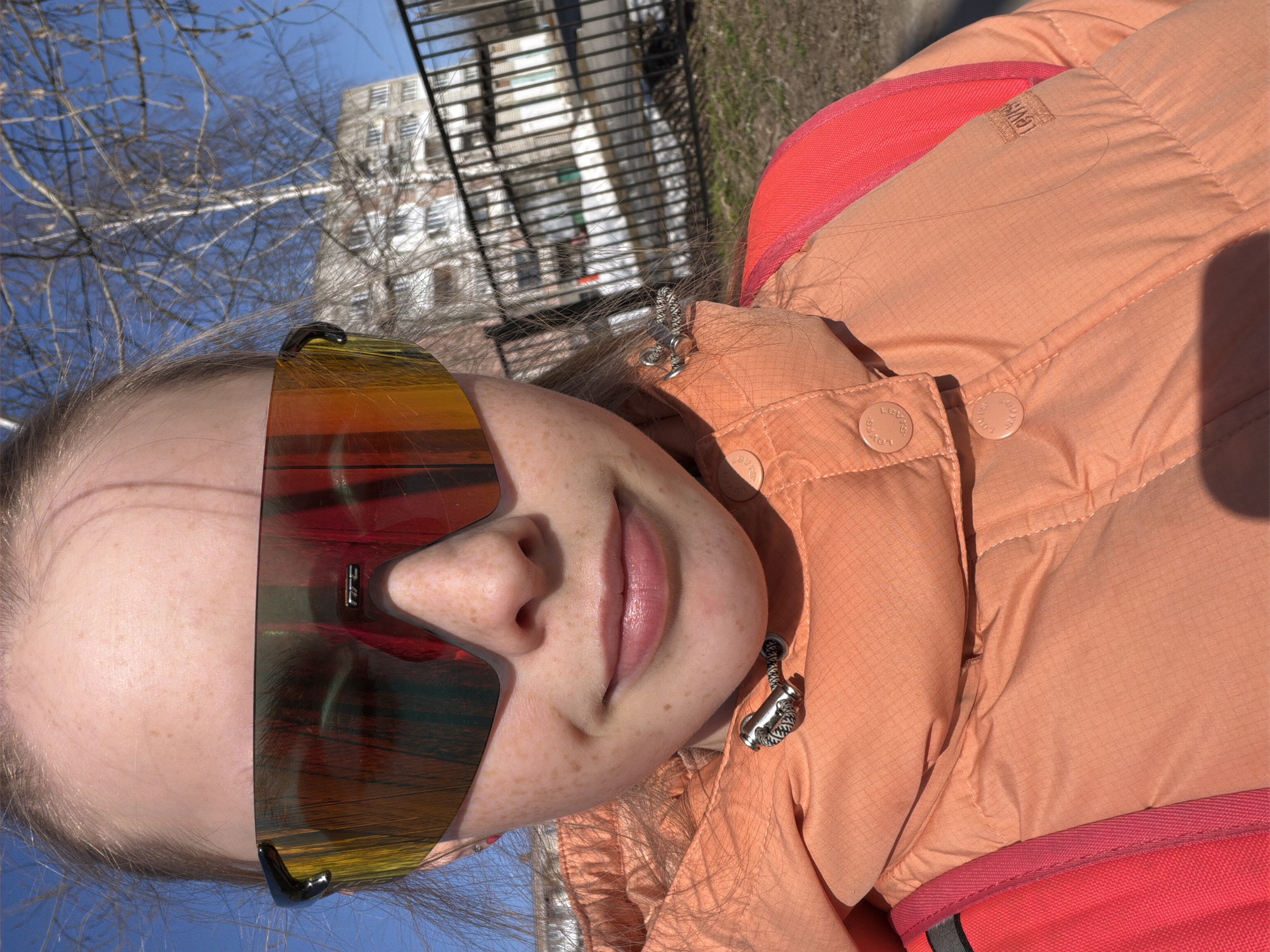 A young, fair-skinned person with freckles, wearing large, reflective sunglasses and a peach-colored jacket with red trim and Levi's branded buttons, is the primary subject of this outdoor, close-up photograph. The image is oriented vertically.
In the background, a blurred cityscape is visible, featuring a tall, light-colored building and a dark, metal fence. Bare branches of trees are also visible against a bright blue sky. The scene appears to be well-lit, possibly on a sunny day.
The location is Berezniki, Russia.