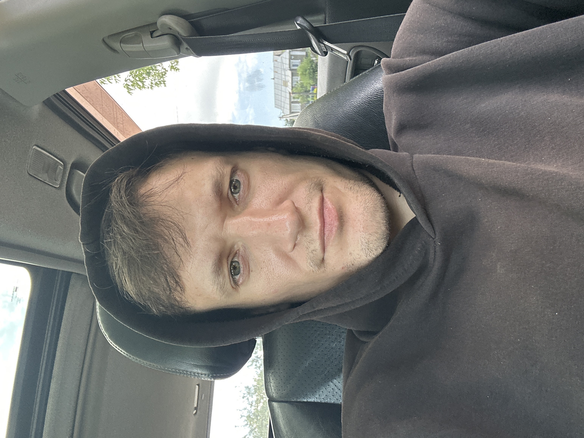 A close-up, selfie-style photograph captures a young Caucasian man with short, messy brown hair and light green eyes, wearing a dark brown hooded sweatshirt. He is seated inside a vehicle, with the interior visible behind him, including a black leather seat and the car's ceiling with a sunroof. Through the car's windows, a rural landscape is visible, featuring houses with tiled roofs and trees, suggesting a location in a village or outskirts. The man's face is illuminated by natural light, and his expression is neutral. The image appears to be taken during daylight. The exact location in Makiv, Ukraine, is not determinable from the visual cues alone, but the setting is consistent with a residential area in that region.