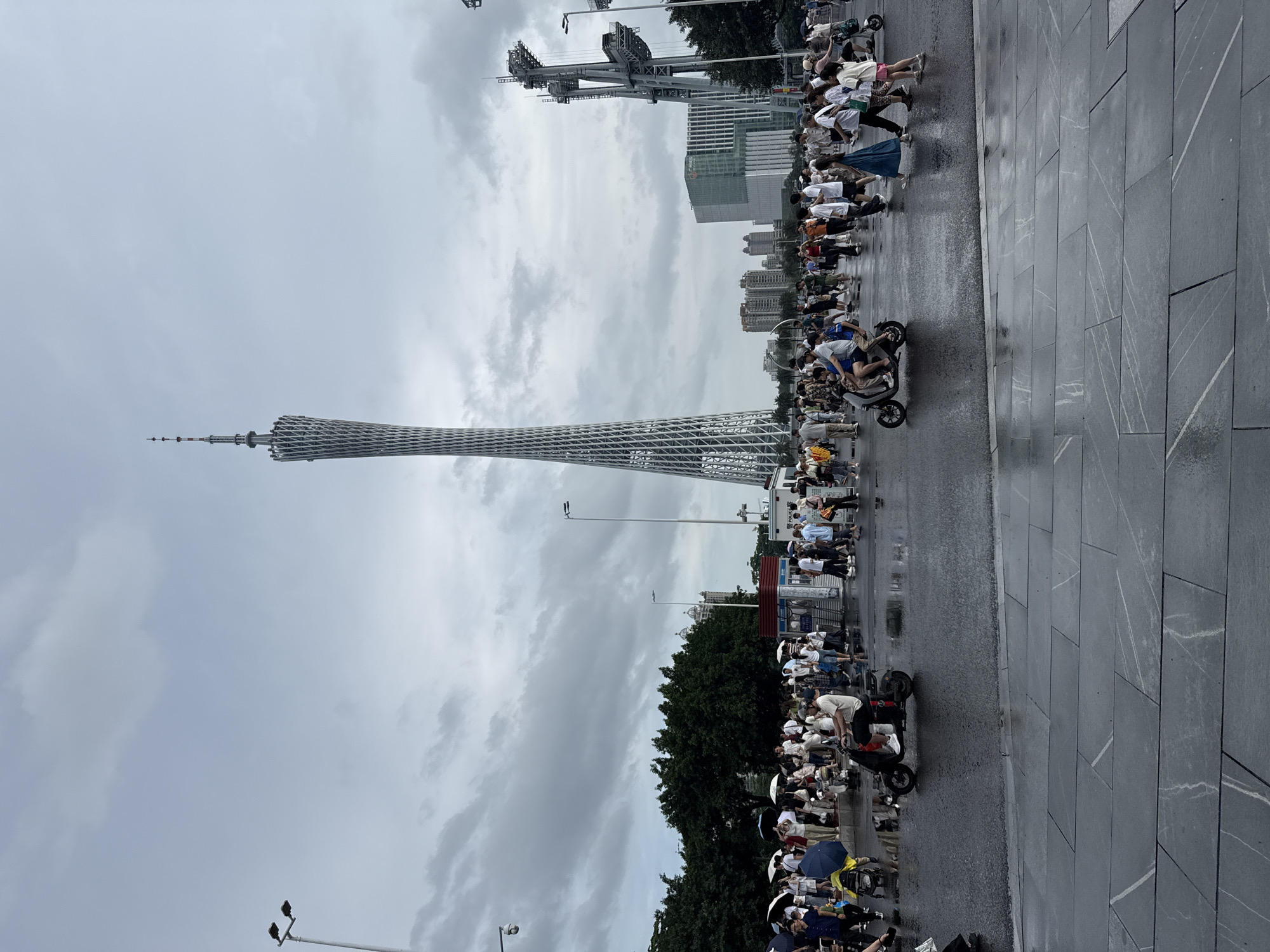 In Tianhe District, China, on a cloudy day, a large crowd of people gathers in an open, paved area with puddles reflecting the overcast sky. Dominating the background is the Canton Tower, a distinctive lattice-like skyscraper, with smaller buildings and structures visible to its right. Several people are riding electric scooters, with one scooter carrying two individuals prominently in the foreground. Many other individuals are walking, some holding umbrellas, indicating a recent or ongoing rain. The ground is wet and reflective, suggesting it has rained or is still raining. Trees line the left side of the scene, and a large, modern structure with a prominent antenna is located to the right of the Canton Tower. A white booth with "POLICE" signage is visible among the crowd.