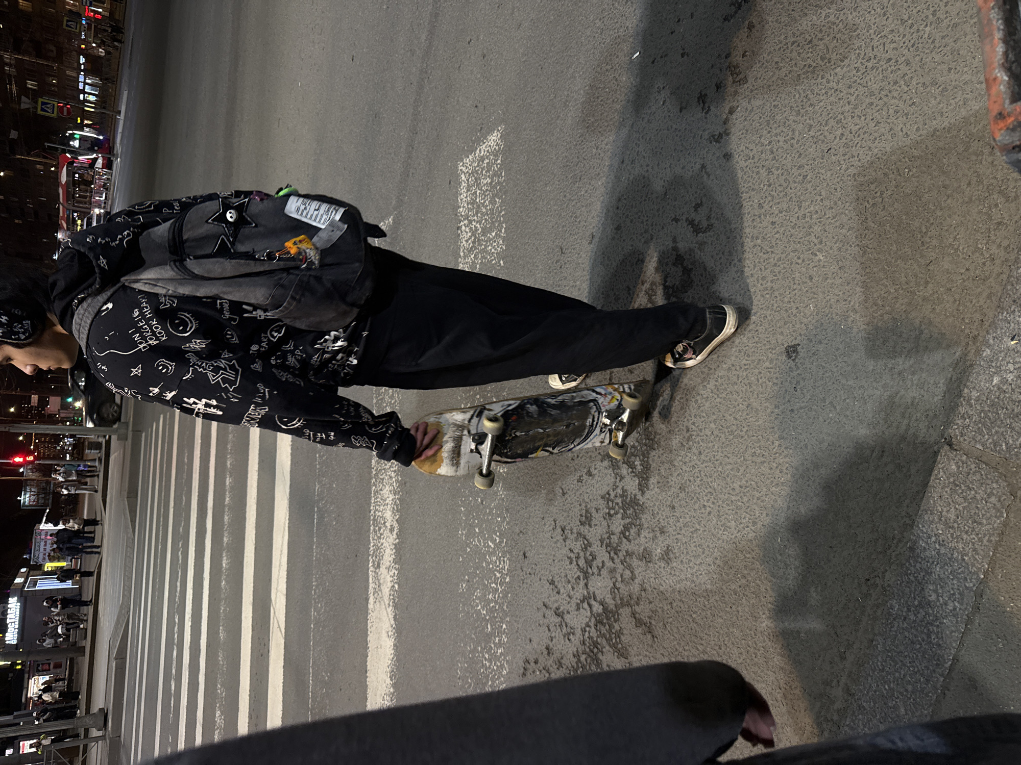 A person stands on a city street at night, holding a skateboard. They are wearing a black hoodie with white designs, black pants, and sneakers. A backpack is slung over their shoulder. The location is Saint Petersburg, Russia, as indicated by the Cyrillic writing on a nearby storefront. The person appears to be standing on or near a crosswalk, evident by the white stripes on the road. In the background, city lights and pedestrian traffic are visible. The environment suggests an urban setting at dusk or night.