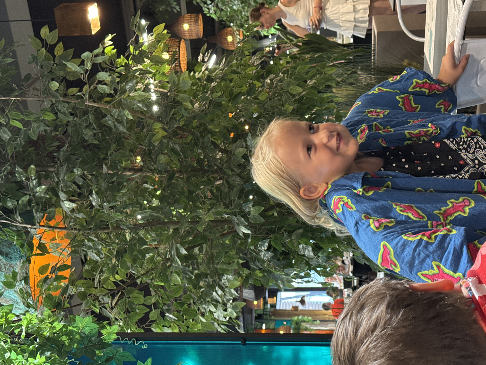 A young girl with blonde hair and a blue shirt with a pink and green pattern is sitting at a table in what appears to be a restaurant. She is smiling and looking to her right. Behind her, a large artificial tree with green leaves dominates the scene. In the background, another woman in a white dress is visible, holding a plate. Several lamps are hanging from the ceiling, and some plants are in planters. The overall impression is of a lively, indoor dining setting.