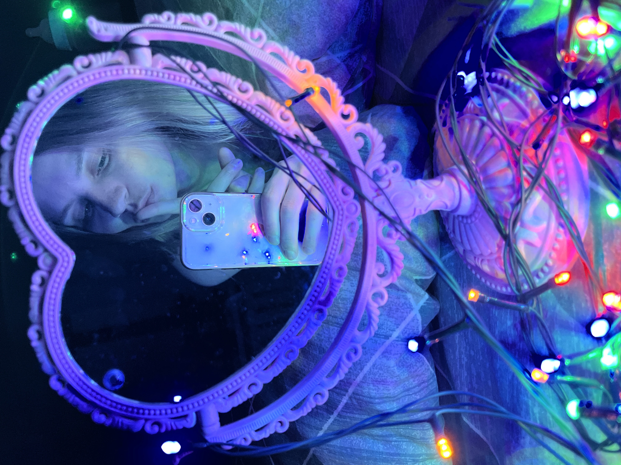 A selfie taken in Chaikovsky, Russia shows a person reflected in a pink heart-shaped mirror, holding a smartphone in front of their face. The mirror is positioned on a pink stand and is surrounded by colorful string lights, creating a vibrant, illuminated effect. The background suggests an interior setting with a patterned fabric or wallpaper. The person in the reflection appears to be looking at their phone.