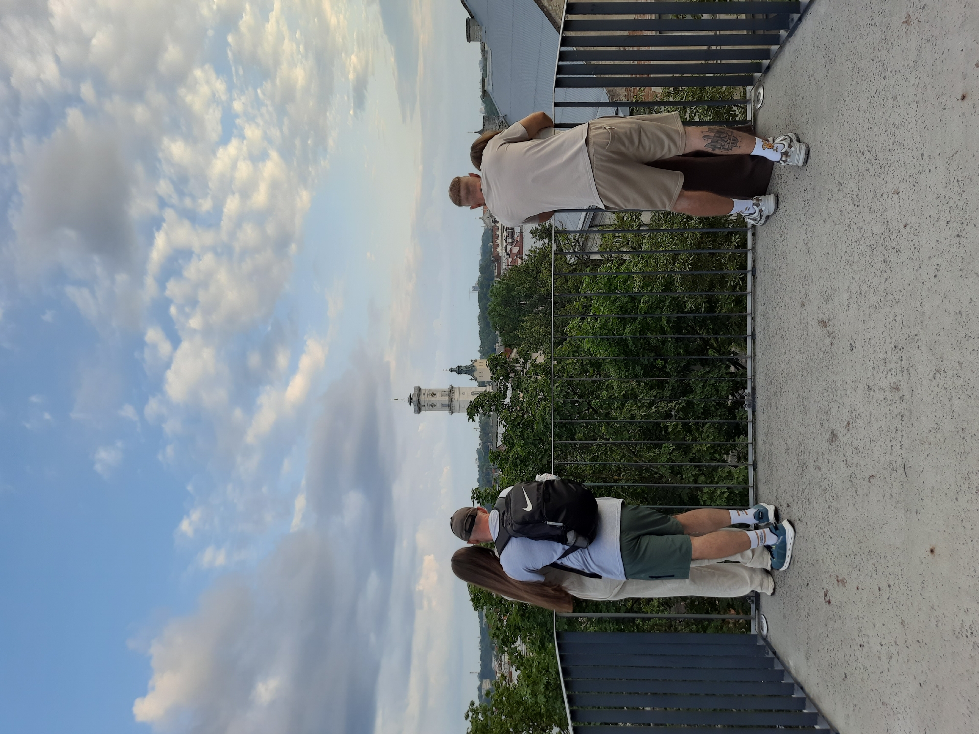 The image depicts two couples on a balcony overlooking the city of Lviv, Ukraine. In the foreground, a man wearing a grey t-shirt, khaki shorts, white socks, and white sneakers, and a woman wearing a brown dress are embracing. To their left, another couple stands. The man wears a grey t-shirt, green shorts, white socks, and blue sneakers, with a black backpack adorned with a Nike logo on his back. The woman wears a light-colored top and light-colored pants.
Behind the railing, lush green trees partially obscure a view of Lviv's cityscape, which features red-tiled roofs and the prominent white clock tower of the Assumption Church (also known as the Latin Cathedral) and a smaller church spire. The sky is partly cloudy with patches of blue and streaks of grey and white clouds. The balcony floor is made of grey concrete.