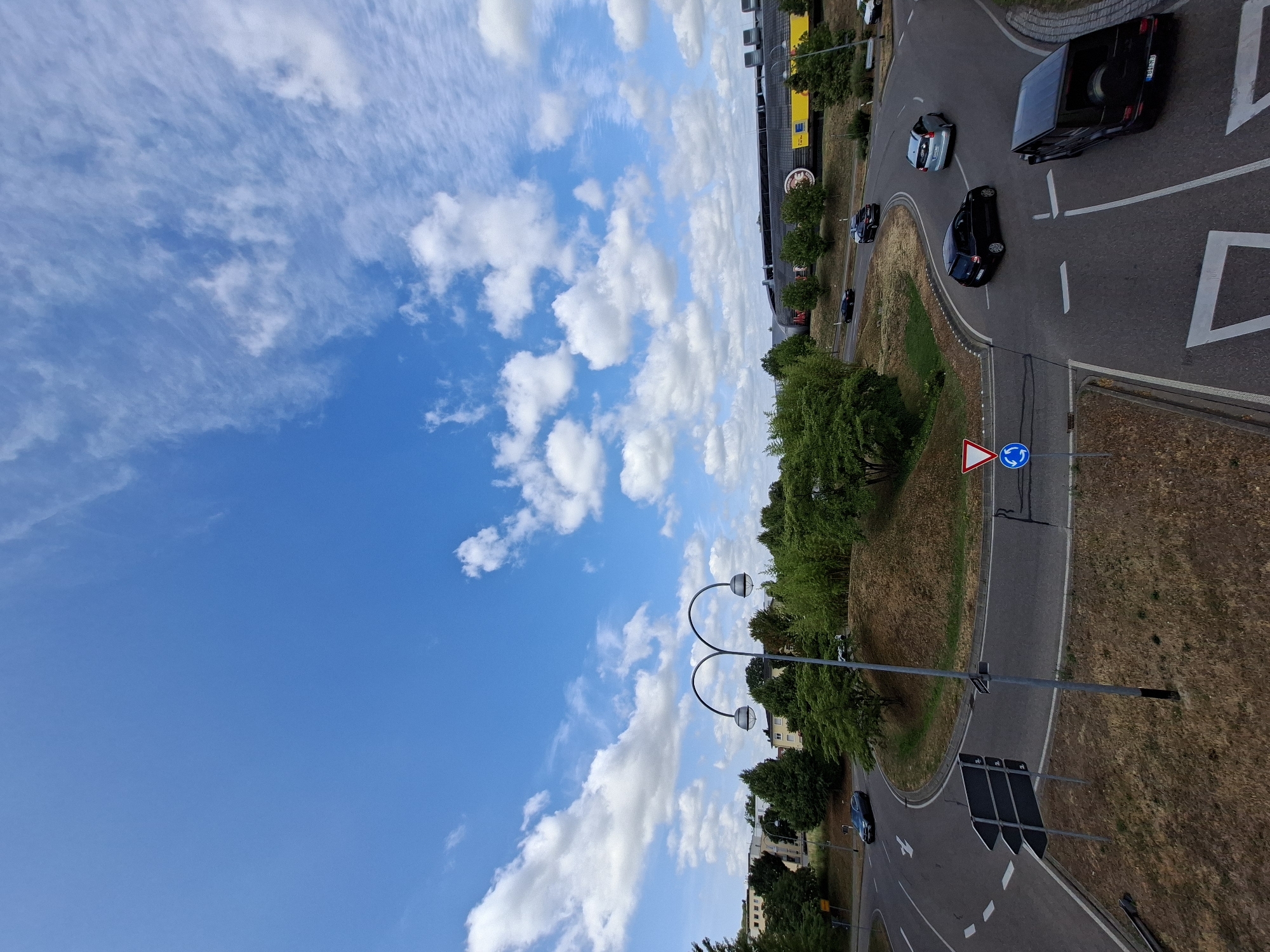 This is a wide-angle, eye-level, daytime, exterior shot of a roundabout with several cars present. The sky is bright blue with scattered white clouds. The roundabout is paved and has lane markings. Several trees and bushes surround the roundabout, with some buildings visible in the background. There are also streetlights and traffic signs present.
Specifically, this image depicts the roundabout at the intersection of Kaiserallee and Lichtentaler Allee in Baden-Baden, Germany. A black Mercedes-Benz G-Class SUV is in the foreground, turning right into the roundabout. To its left, a black hatchback is entering the roundabout from the same direction. Further around the roundabout, a silver hatchback is visible, and a dark sedan is exiting the roundabout. In the background, a large building with yellow signage, likely a commercial establishment, is visible. The overall environment is suburban, with a mix of greenery and urban infrastructure.