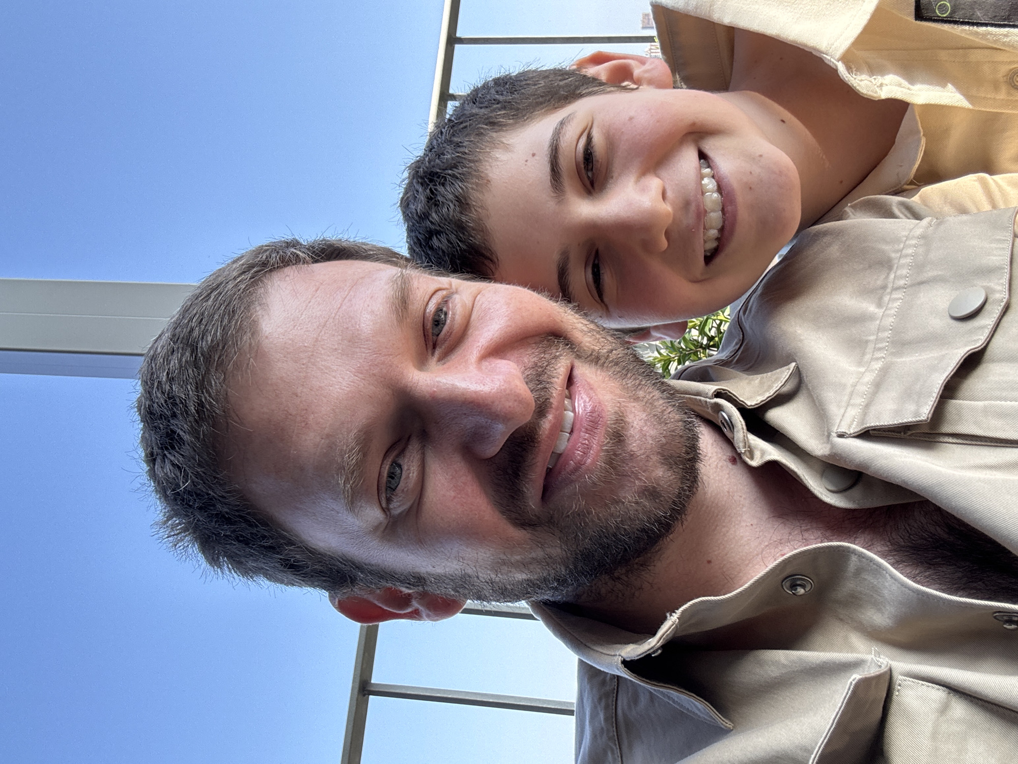 Two individuals, an adult male and a younger male, are in close proximity, possibly posing for a selfie-style photograph. The adult male has short, receding dark hair, a beard with graying hairs, and is wearing a tan button-up shirt or jacket. The younger male, likely a child, is positioned slightly behind and above the adult, with a visible smile revealing braces. Both are looking toward the camera. The background includes a clear blue sky, metal railing, and hints of greenery, possibly indicating a balcony setting. The location appears to be urban, with potential glimpses of buildings. Based on the architectural elements and visual cues, a tentative location of Milan, Italy can be suggested, though definitive confirmation would require more specific landmarks or contextual clues.