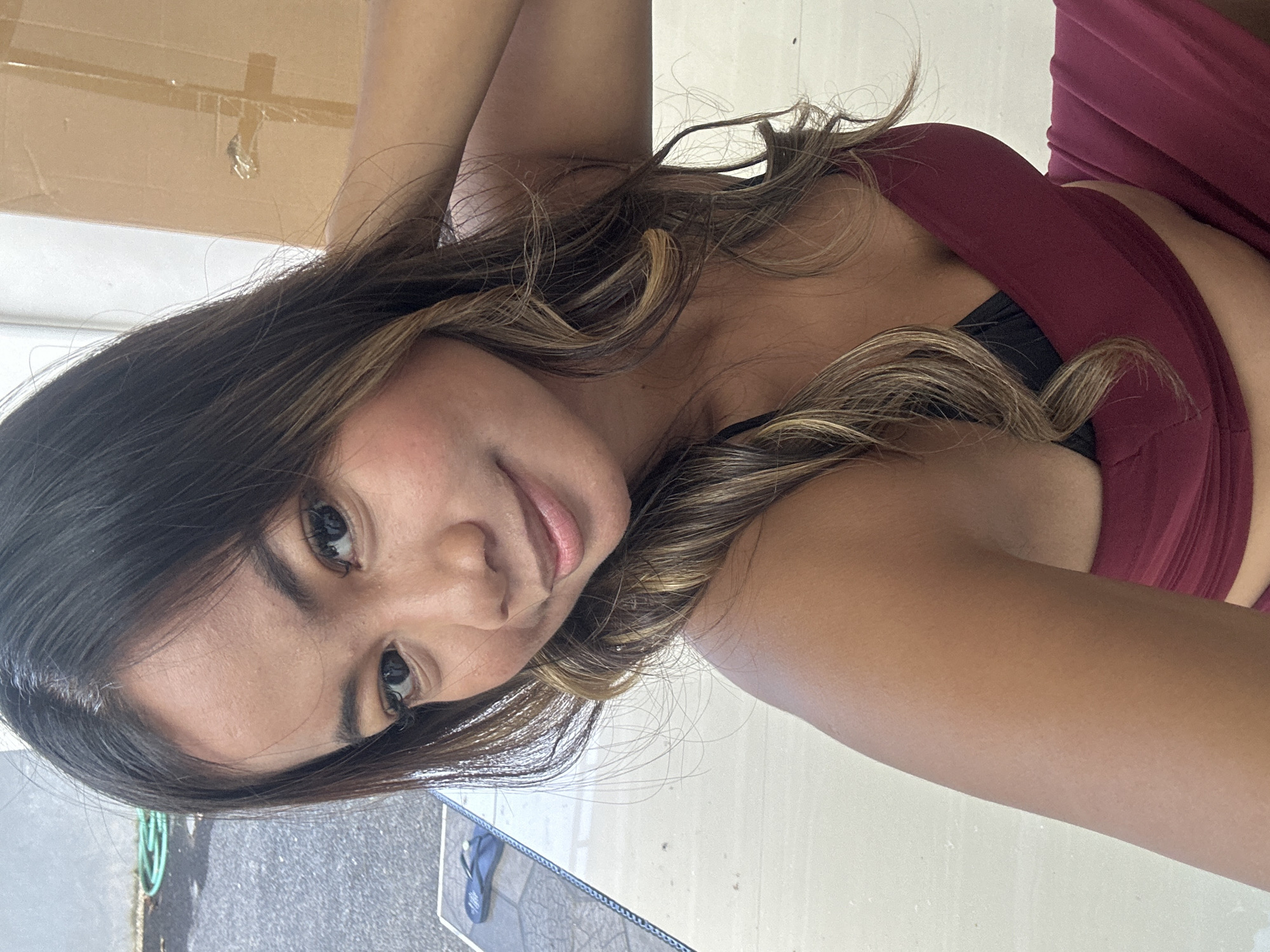 A young woman with long, wavy brown hair with blonde highlights smiles at the camera. She is wearing a maroon top over a black bra and a maroon skirt. She is leaning on a white table with a black edge, and a pair of black flip-flops are visible on the table to her left. In the background, a grey wall and a brown cardboard box are visible, along with some greenery and a concrete surface. The image appears to have been taken outdoors during the daytime, possibly in Sukawati, Indonesia.
