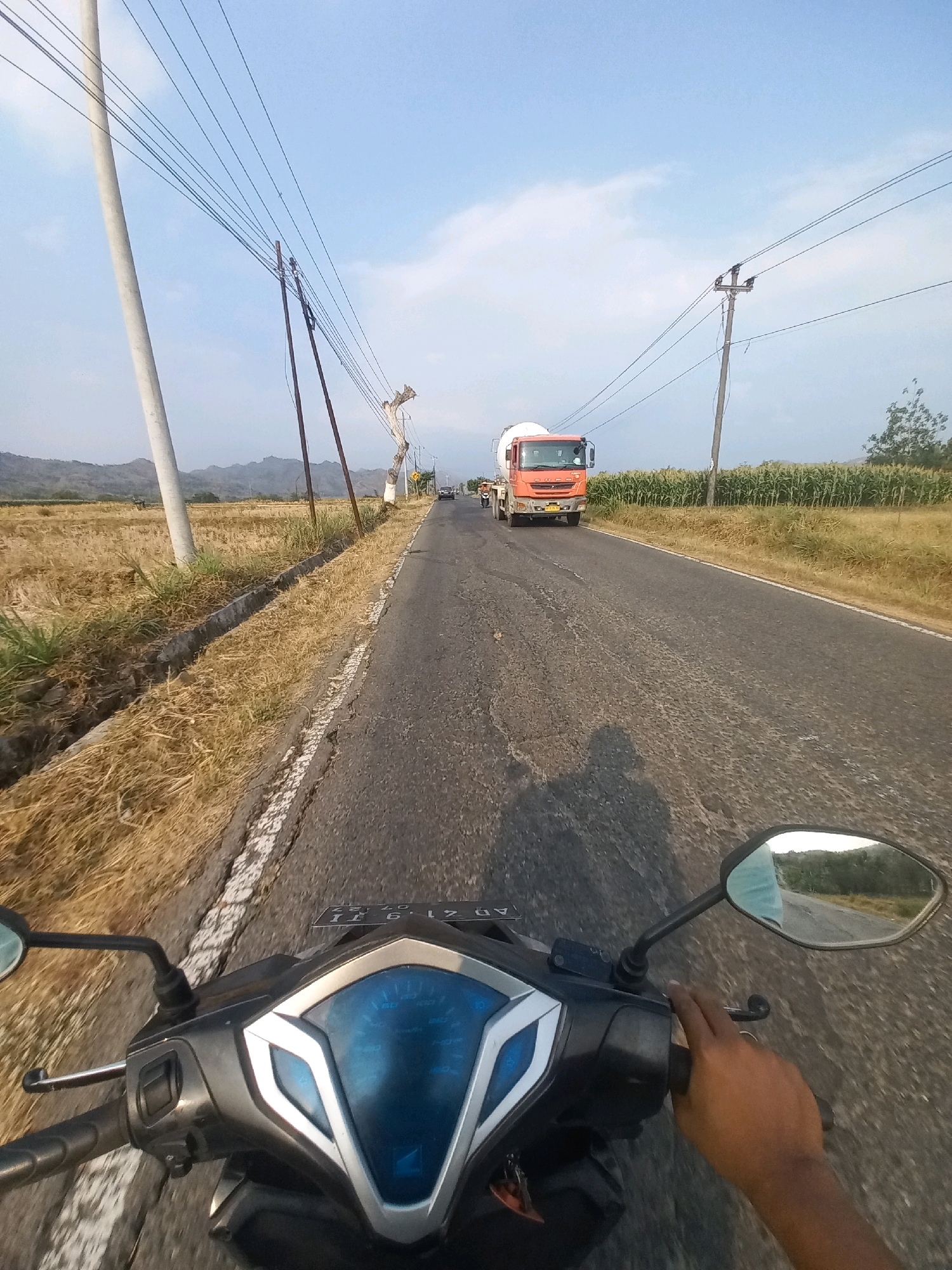 The image is a first-person perspective shot from a motorcycle traveling on a paved road in Baturetno, Indonesia. The road surface is worn with patches of rough texture and a broken white line demarcates the lane. On the left side of the road, dry, yellow grass and a concrete drainage ditch line the embankment. Beyond the embankment, a field of dry vegetation stretches towards a distant hilly landscape. On the right side of the road, a field of green corn stalks grows. Overhead, multiple power lines are strung between utility poles, with one concrete pole on the left and a wooden pole on the right supporting the lines. In the distance, an orange cement mixer truck is visible on the road, approaching the motorcycle. The sky is a clear blue with a few scattered white clouds. The motorcycle's dashboard with its blue digital display is prominently in the foreground, and a rider's hand is visible gripping the handlebar. A shadow of the rider and motorcycle is cast on the road. A side mirror on the right shows a reflection of the sky and distant hills.