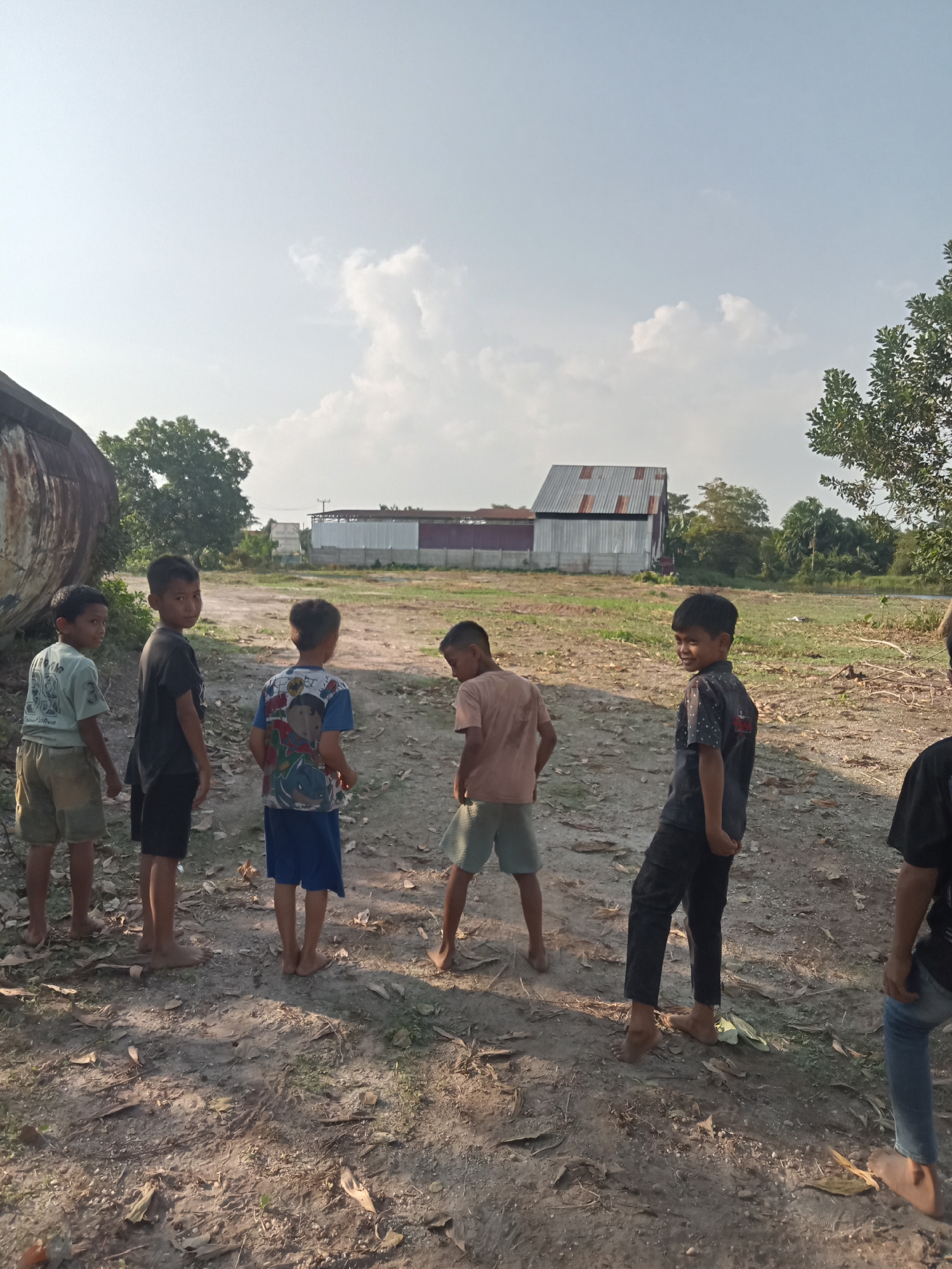 In Teluk Kenidai, Indonesia, six boys, aged approximately 7-12 years old, stand on a dirt path. The boys are dressed in casual clothing, including t-shirts and shorts. They are facing in various directions, with some looking at the camera and others looking away. In the background, there is a large, cylindrical object on the left, likely a storage tank. Further in the background, there are buildings with corrugated metal roofs and trees. The environment appears to be an open, rural area with dry vegetation and debris on the ground. The sky is clear with a few scattered clouds. The lighting suggests it is daytime, possibly late afternoon, given the shadows. The overall scene depicts a group of children in an outdoor setting.