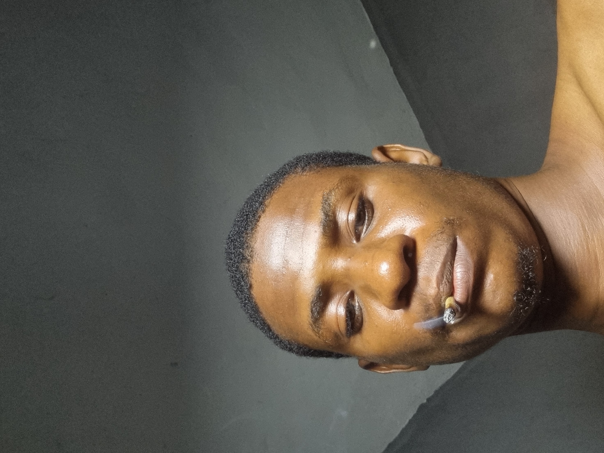 The image is a close-up, head-and-shoulders selfie of a young Black male. He has short, curly dark hair and dark brown eyes. He is looking down and slightly to the left. His lips are parted, and he is holding a lit cigarette in his mouth. Smoke is visible rising from the cigarette. He has some stubble on his chin and upper lip. The background is a dimly lit, textured wall. The lighting suggests an indoor setting, possibly at night or in a room with limited light sources. There is no specific indication of the location being Nkpor, Nigeria; this is an assumption.