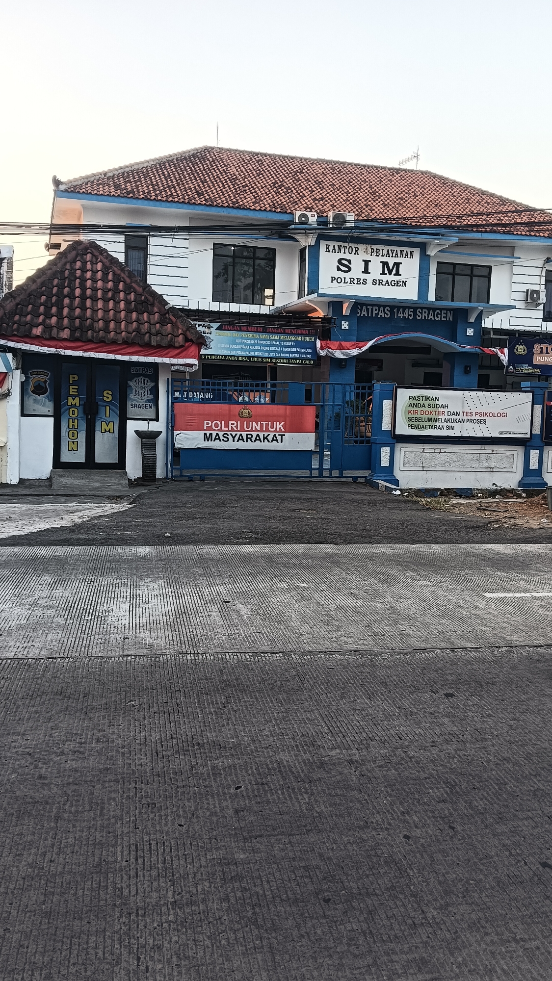 This is an exterior, medium-angle shot of the Satpas 1445 Sragen building, a police station in Sragen, Indonesia. The building is two stories tall with a white facade and a brown tiled roof. A blue awning with the words "KANTOR PELAYANAN SIM POLRES SRAGEN" is visible above the main entrance. In front of the building is a blue gate with a red and white banner that reads "POLRI UNTUK MASYARAKAT." To the left of the gate is a smaller building with a brown tiled roof and a sign that reads "PEMOHON SIM." The ground in front of the building is paved with concrete and shows wear and tear. The overall scene appears to be a public service building with clear signage indicating its purpose.