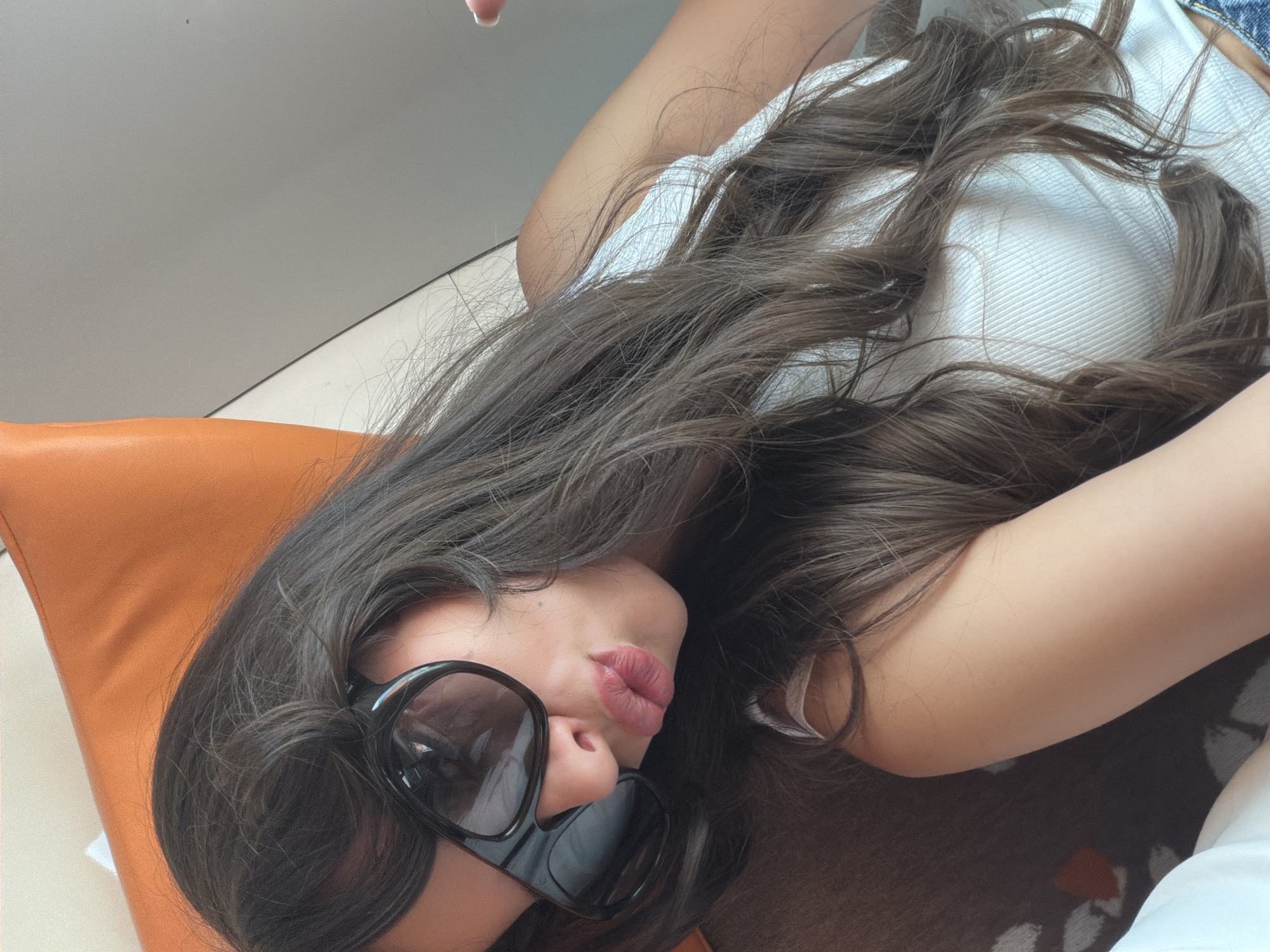 The image presents a close-up, tilted view of a person, likely female, reclining indoors. The subject's head is angled to the left, adorned with long, dark brown, wavy hair that flows across her body. She wears prominent, dark-lensed sunglasses that cover her eyes. Her lips are a focal point, colored with a pink or red lipstick and pursed in a distinct pout.
She is dressed in a light-colored, possibly white, ribbed top. A hint of blue denim fabric is visible in the upper right corner of the frame, suggesting she may be wearing jeans or shorts. To the left, an orange cushion or pillow adds a vibrant color element to the composition. The background largely consists of a plain, light gray or white surface, possibly a wall or ceiling, with a discernible seam. The person's right arm is partially visible in the lower right, resting on a dark surface with a subtle pattern, which could be a rug or upholstery.
The lighting appears soft and diffused, consistent with an indoor setting, creating a relaxed atmosphere. The image focuses on the subject's expression and style. No text is visible in the frame. The provided context indicates the location is Cagnes-sur-Mer, France.