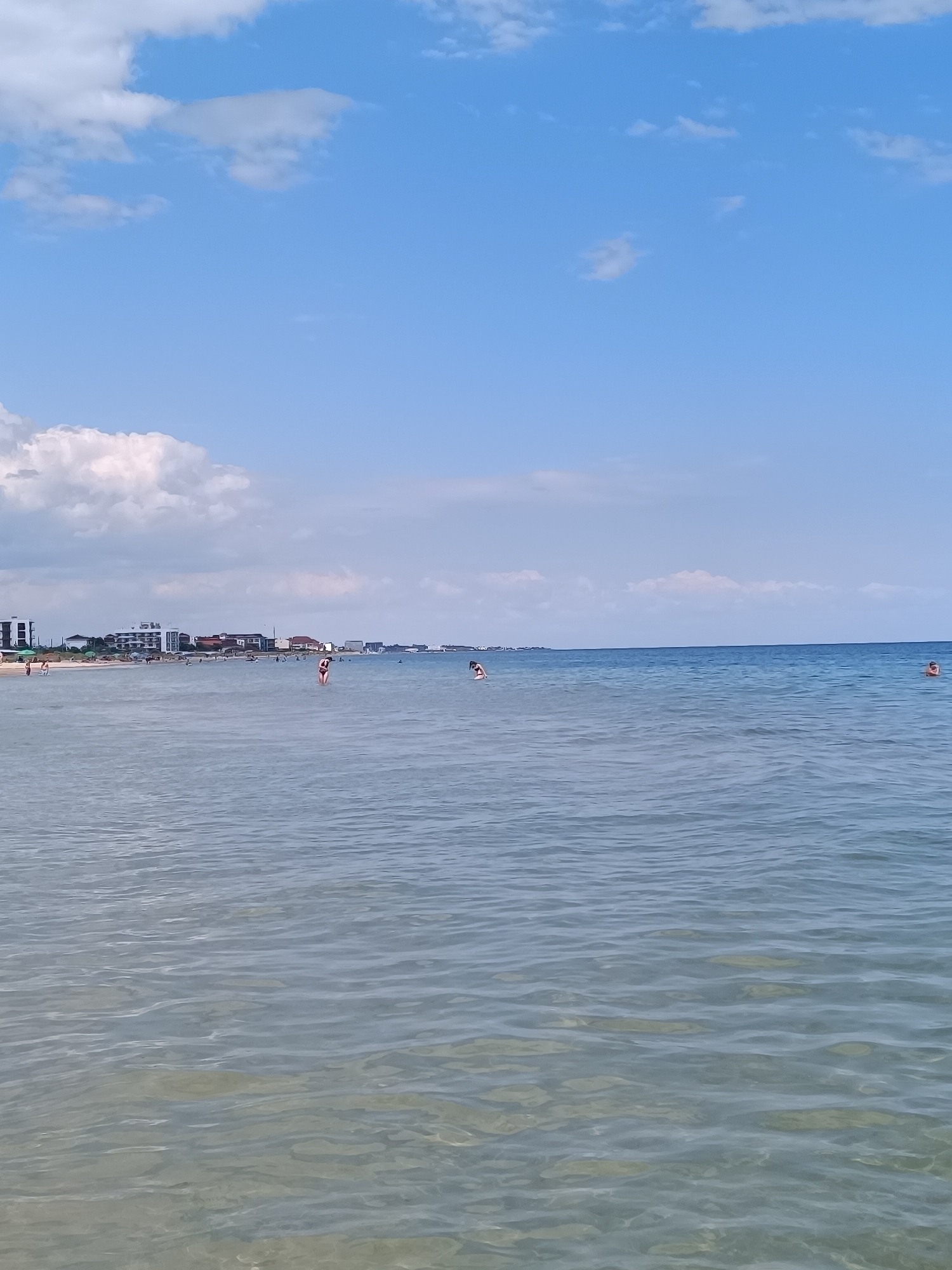 This is a full shot of a beach in Zatoka, Ukraine. The sky is blue with scattered white clouds. The sea is calm and clear with gentle waves. Several people are visible in the water and on the beach. In the foreground, the clear water allows a view of the sandy bottom. Further out, a few people are wading or swimming. In the mid-ground, the shoreline is lined with buildings, some appearing to be hotels or apartment complexes, along with umbrellas and beachgoers. The overall scene depicts a typical summer day at a seaside resort.