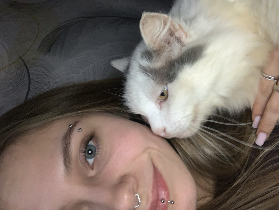 The image depicts an indoor setting, consistent with a residence in the city of Foki, Russia. A female subject, primarily seen in a close-up, occupies the left side of the frame. She has fair skin, blue eyes, and long, light brown hair. Her left eyebrow features two silver ball piercings. A silver hoop with small white stones is visible in her septum, and her lower lip has a vertical labret piercing with two silver ball studs. She exhibits a subtle smile, with her gaze directed towards the camera.
She is holding a domestic cat, positioned to the right of her face. The cat is predominantly white with gray fur patches on its forehead and right ear, and possesses light yellow/amber eyes with prominent white whiskers. The cat's head is nuzzled against the female's left cheek, and its gaze is directed slightly upwards and to the right. The female's right hand, with light pink painted fingernails and two silver rings on her ring finger, is visible gently supporting the cat's underside. The background consists of a dark gray wall with a subtle, textured feather-like pattern.