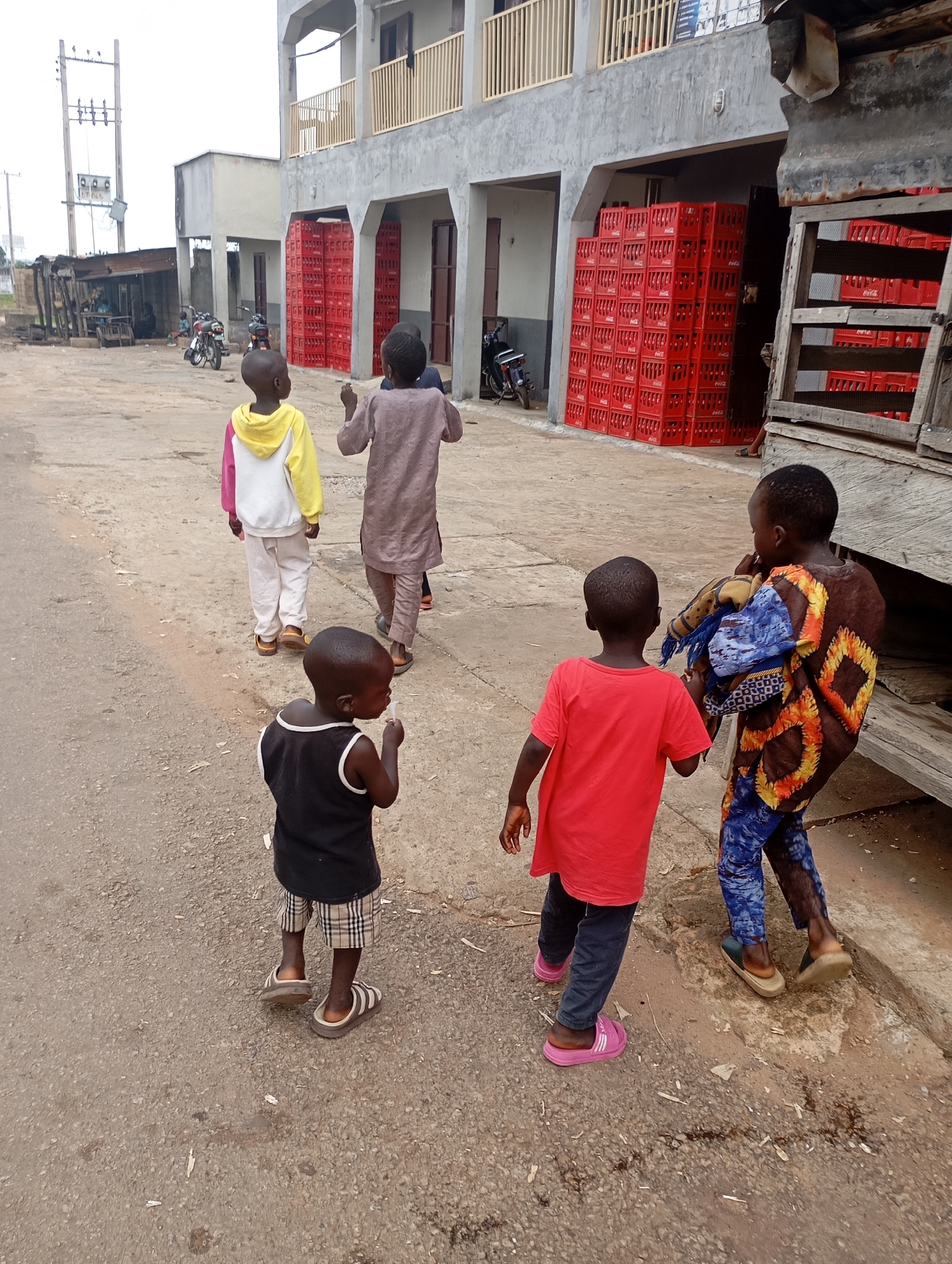 A group of six children walk along a dusty road in Shendam, Nigeria. In the foreground, a young boy in a black tank top and plaid shorts chews on something white. Behind him, a child in a red t-shirt and dark pants walks with another child wearing a patterned blue and orange shirt and blue pants. Further down the road, a child in a yellow and pink hoodie and white pants walks with two other children, one in a brown tunic. In the background, red crates are stacked high outside a building with a balcony. Motorcycles are parked near a small kiosk with a corrugated metal roof. Overhead, power lines and poles are visible against a cloudy sky. The scene appears to be an outdoor marketplace or commercial area.