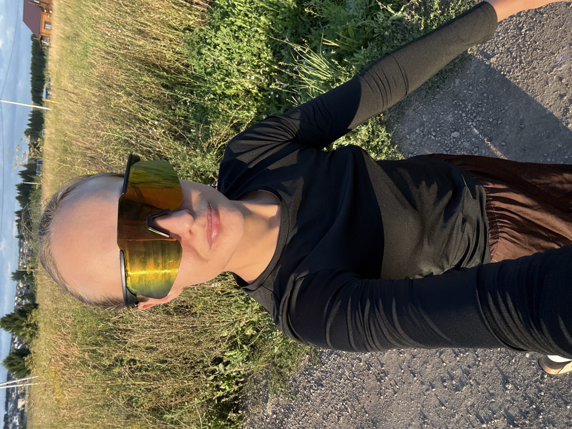 A woman is taking a selfie outdoors in a rural area, likely Podshivalovo, Russia. She is wearing a black long-sleeved shirt, brown pants, and large, mirrored sunglasses. Her hair is pulled back, and she has light makeup on. The setting appears to be a grassy field next to a gravel path, with trees and buildings visible in the background. The sun is low in the sky, casting long shadows, indicating it is either early morning or late afternoon. The weather appears to be clear and sunny. The woman's expression is neutral. There is no visible text in the image.