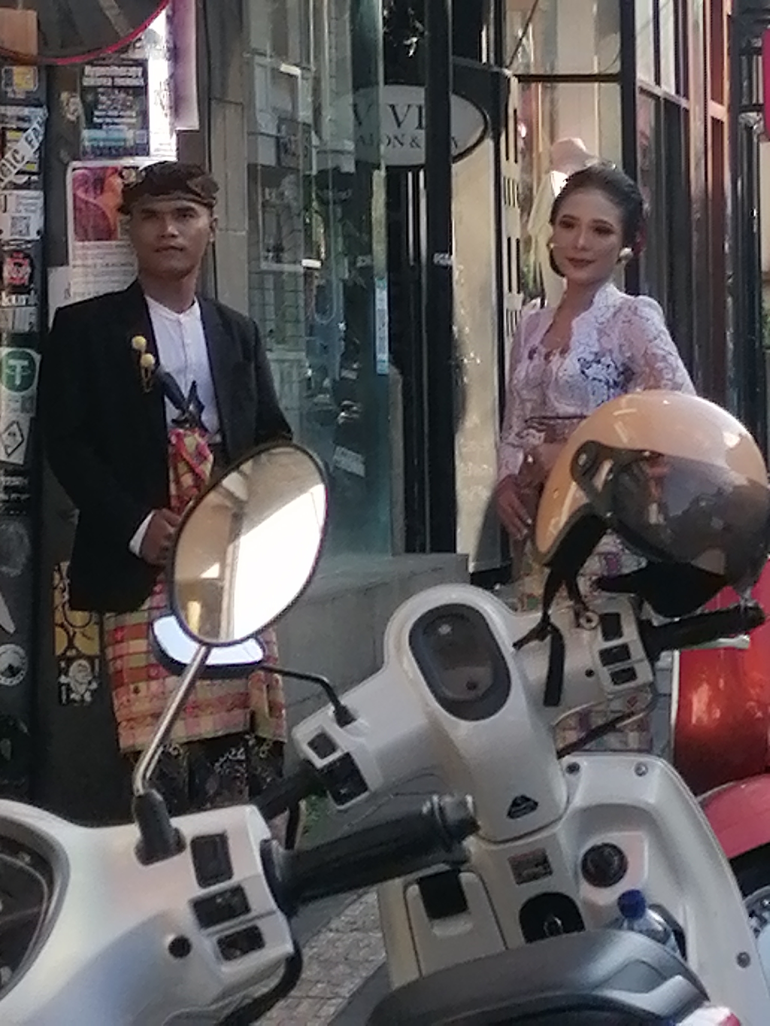 Two individuals are standing outside a shop. The person on the left, a male, is wearing a black jacket over a white collared shirt and a patterned sarong. He has dark hair and is looking forward. The person on the right, a female, is wearing a lace top and a patterned sarong. She has dark hair styled up and is looking to her right. Both individuals appear to be dressed in traditional attire. In the foreground, a scooter is partially visible with a helmet resting on its handlebars. The background shows the entrance to a shop with a glass facade, adorned with posters. The scene is likely outdoors in a city environment, possibly in Ubud District, Indonesia, given the attire.