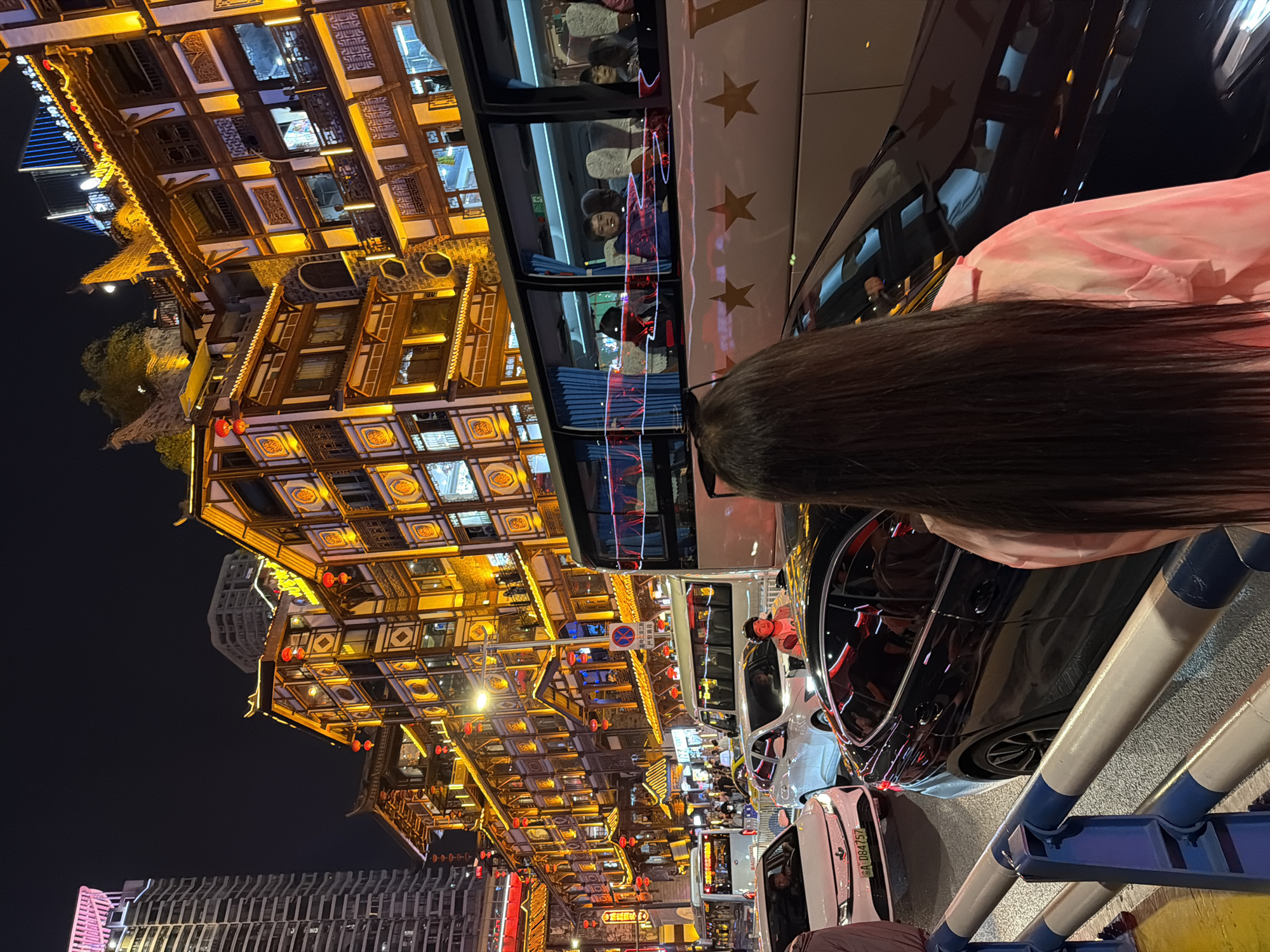 The image captures a vibrant nighttime street scene in Yuzhong District, China, showcasing a blend of traditional and modern urban aesthetics. In the immediate foreground, the back of a person with long, dark hair and a pink garment is visible, positioned behind blue and white striped barriers.
The midground reveals a bustling street with several vehicles. A large white bus adorned with yellow star decals is prominent, its windows reflecting the bright city lights. Inside, several passengers are visible, some smiling, suggesting a tour or commute. Behind the bus is a sleek black car, also reflecting the intense illumination from the surroundings. A white car with a partially visible license plate, "A-084757," is also present. A red circular traffic sign with Chinese characters and a white horizontal bar indicates a prohibition, likely no parking or stopping.
The left side of the frame is dominated by magnificent multi-story traditional Chinese buildings. These structures are intricately designed with ornate wooden frameworks, patterned windows, and multiple tiers. They are brilliantly illuminated with warm, golden lights, and numerous red lanterns hang from their eaves, creating a festive and historic atmosphere. In contrast, modern high-rise buildings with contemporary facades can be seen in the far distance, providing a striking juxtaposition. The scene depicts a lively urban night, filled with architectural beauty and active street life.