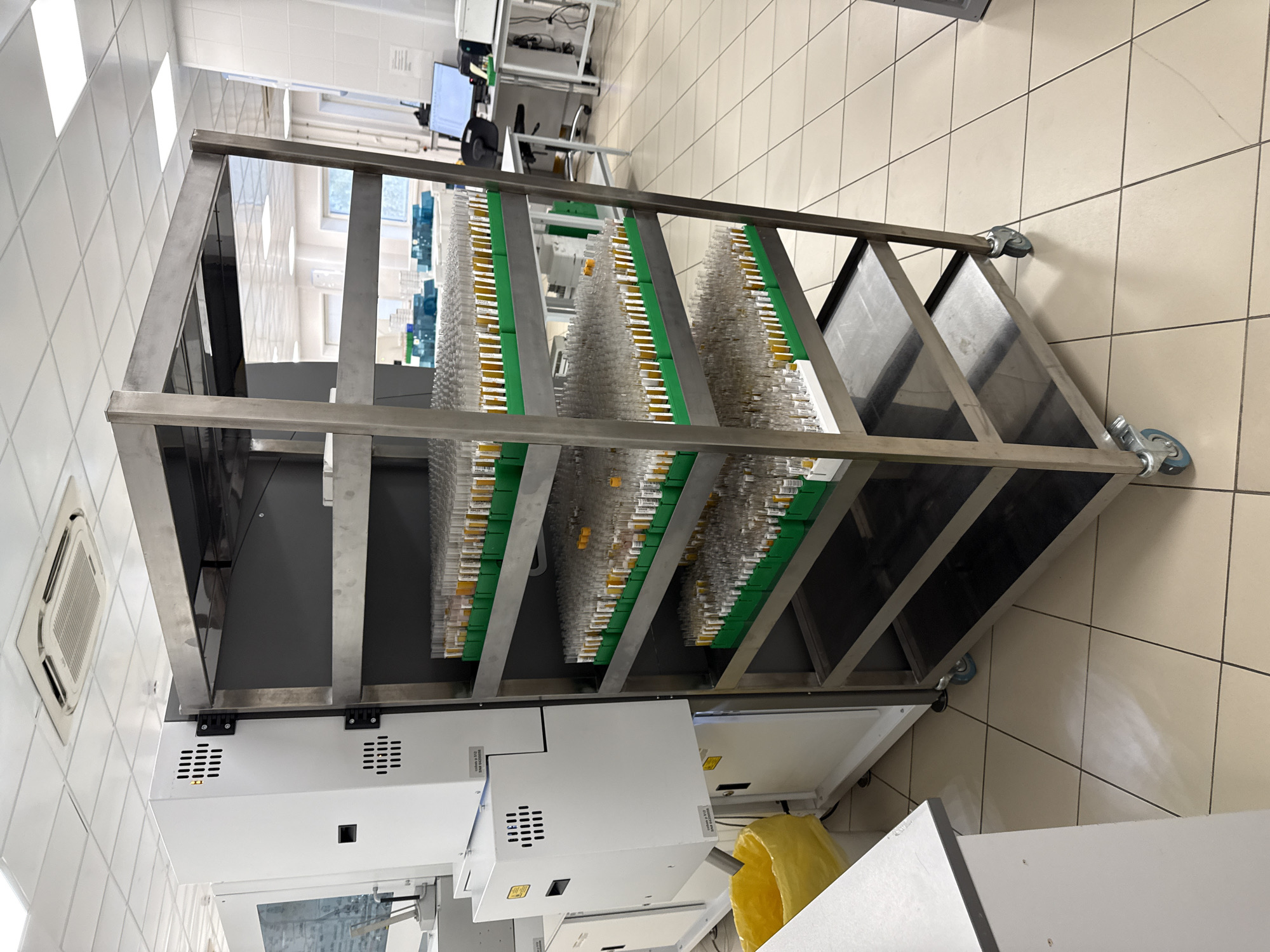 The image shows a metal cart holding multiple racks of test tubes with yellow tops in a sterile environment. The cart has wheels and is placed in a room with tiled walls and floors, suggesting a laboratory or medical facility. A machine with white panels is visible at the bottom of the cart. Based on the equipment and sterile environment, the image likely depicts a medical or research laboratory. There is no information in the image to identify this location as Moscow, Russia.