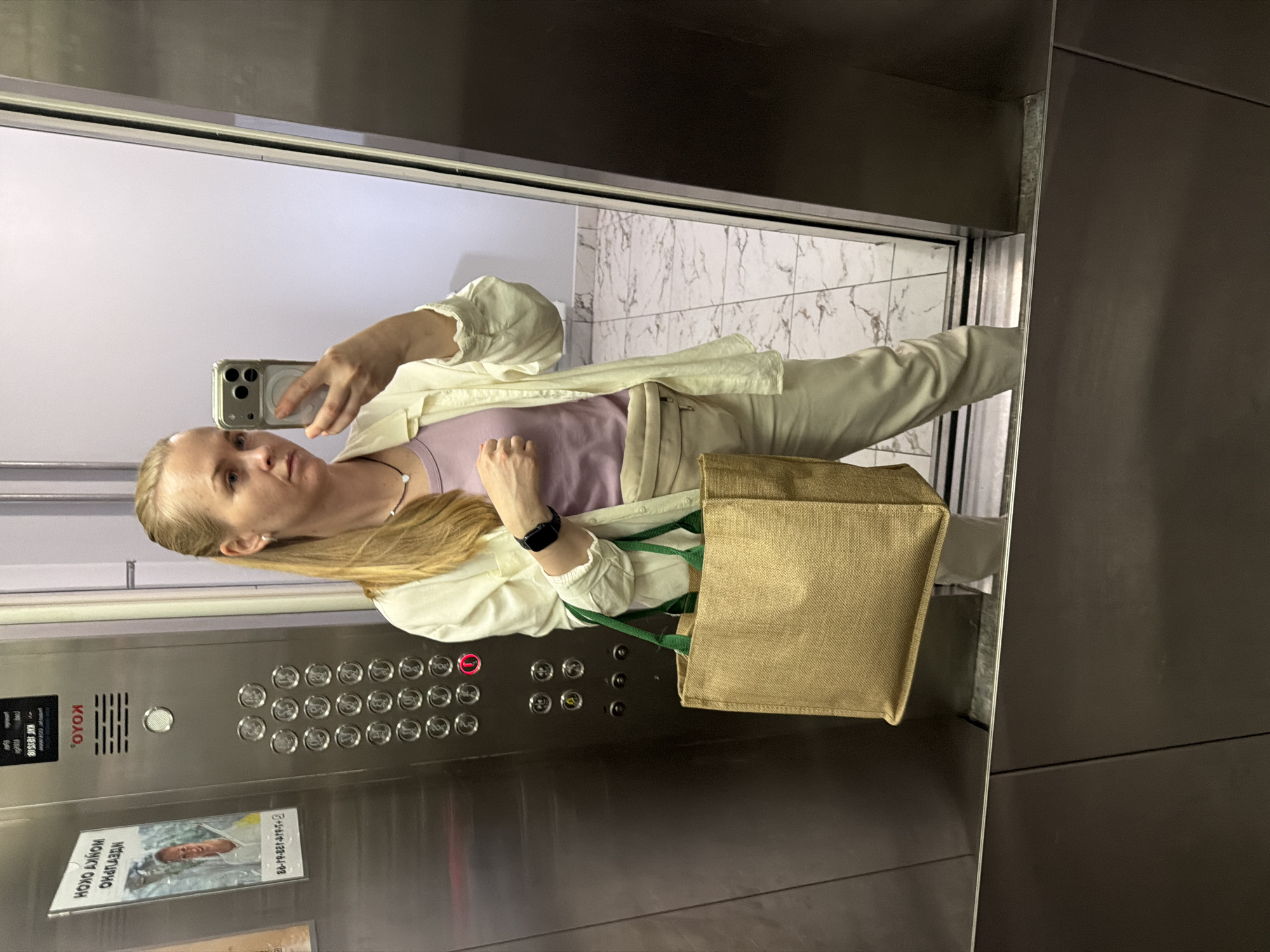 A woman is taking a mirror selfie in an elevator. She is wearing a light-colored unbuttoned shirt over a lavender tank top, and light-colored pants. She has blonde hair pulled back and is wearing a necklace and a watch. She is holding a large, tan, canvas tote bag with green handles in her left arm. The elevator control panel is visible to her left, with a red illuminated button. The floor of the elevator is visible, as is a reflection of the woman and her surroundings on the elevator doors. The image also shows a poster attached to the elevator wall with Russian text and a picture of a smiling person. The poster is located on the left side of the elevator. The elevator is likely located in a residential building or office building in the city of Khabarovsk, Russia, as indicated by the Russian text on the poster and elevator panel.