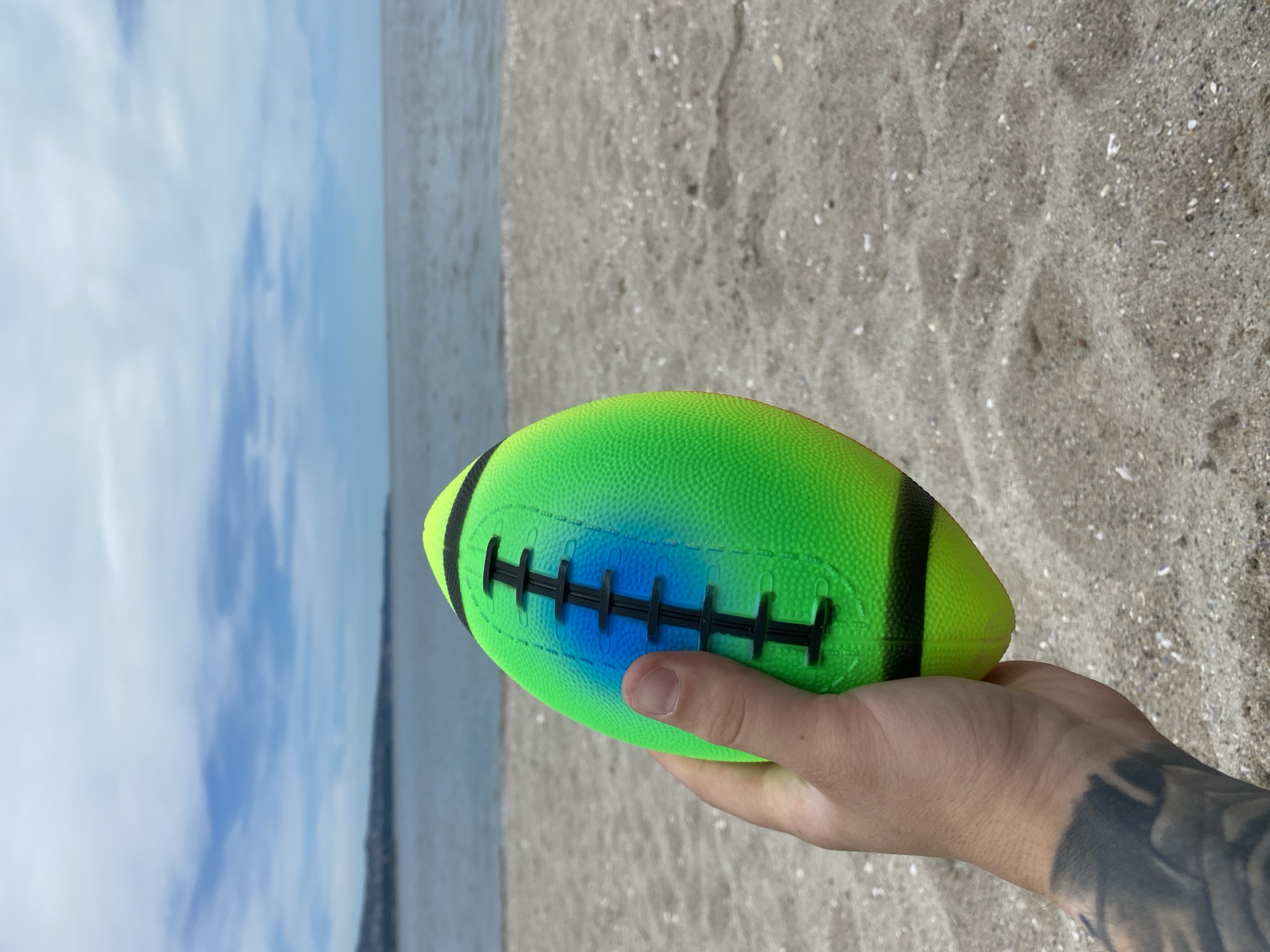 In the city of Varna, Bulgaria, a person's hand, featuring a tattoo on the forearm, holds a vibrant, neon green and blue-hued American football on a sandy beach. The background consists of the calm, greyish-blue sea meeting a hazy horizon with a distant coastline under a cloudy sky. The sand is a light beige color, interspersed with small white shells. The image is taken from a close-up perspective of the football and the hand.