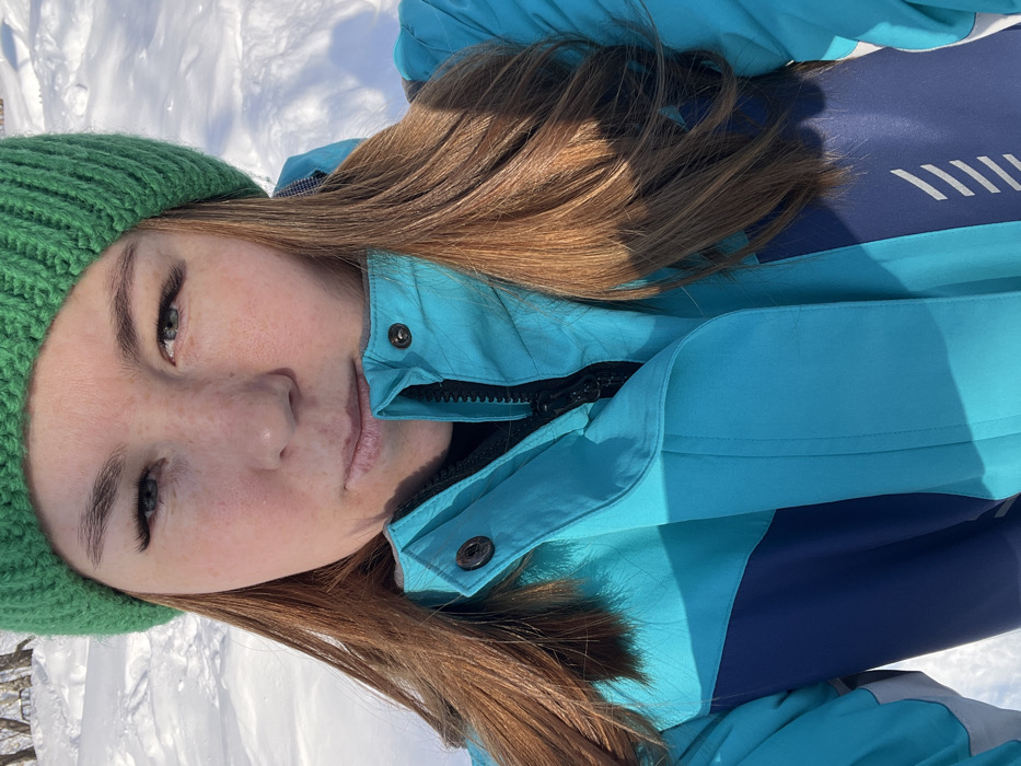 A close-up, chest-up portrait shows a female subject outdoors in a snowy environment within the city of Chaikovsky, Russia. The subject has fair skin with visible freckles, blue-grey eyes featuring eyelash extensions, and long, reddish-brown hair styled down, partially visible beneath a bright green ribbed knit beanie. She is wearing a multi-colored winter jacket, predominantly turquoise with navy blue sections on the torso and sleeves. The jacket's main closure is a black zipper, which is partially open, revealing a dark inner layer. Two black snaps are visible on the collar flap of the jacket. The subject's gaze is directed towards the camera with a neutral expression. The background consists of a brightly illuminated, deep snow cover, with faint outlines of bare trees in the distance under clear, sunny conditions.