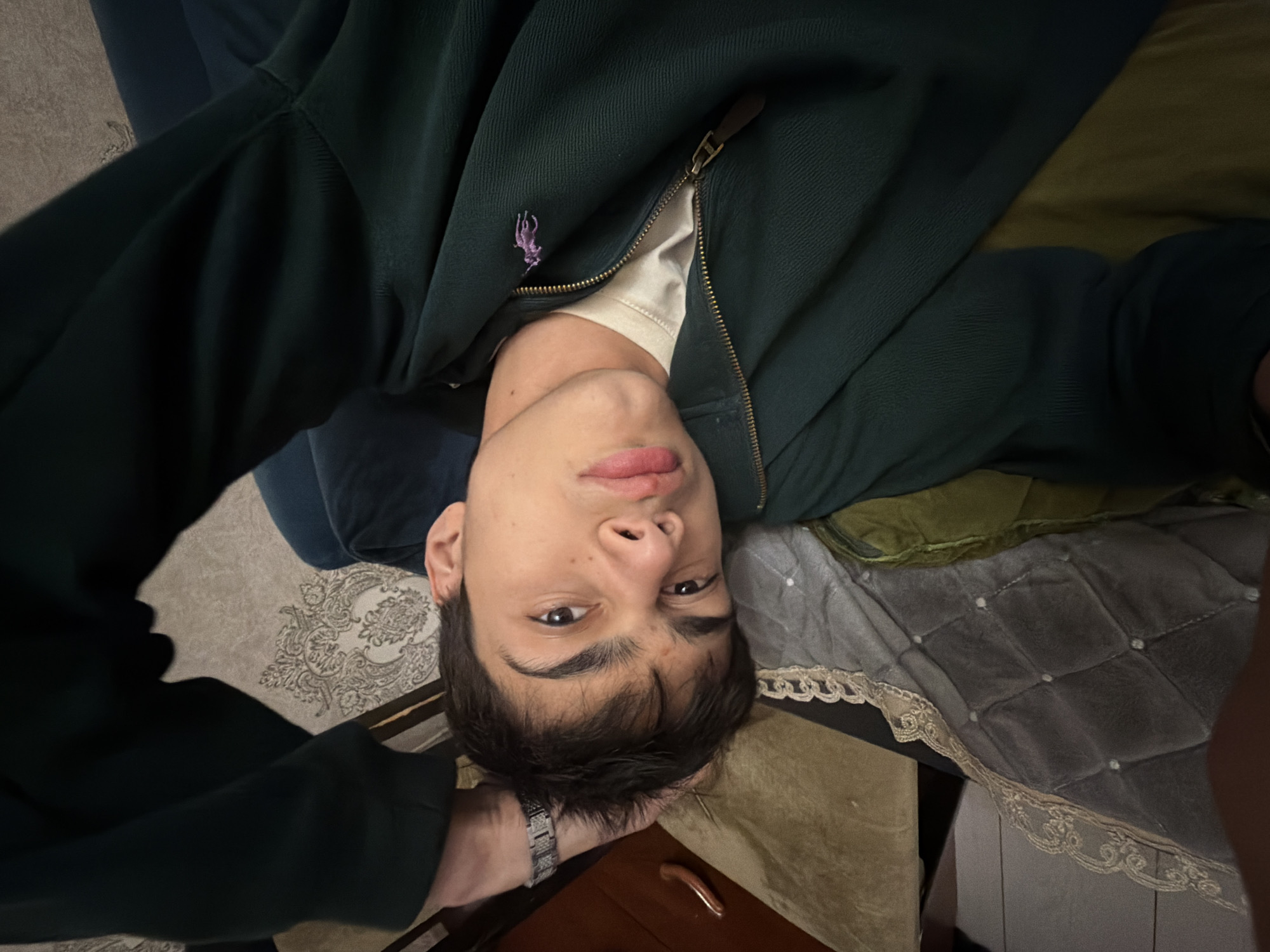 The image shows a close-up, angled shot of a young person with dark hair lying down on a bed or sofa. They are wearing a dark green zippered jacket over a light-colored t-shirt, and a silver watch is visible on their left wrist. The person's head is resting on a patterned pillow, and a part of a blanket or throw with decorative trim is visible in the foreground. The background includes a wall with a floral wallpaper pattern and a dark blue surface, possibly furniture. The lighting is dim, and the overall impression is of a casual, indoor setting. No specific details about the location in the city of Unknown, Russia are discernible from the image.