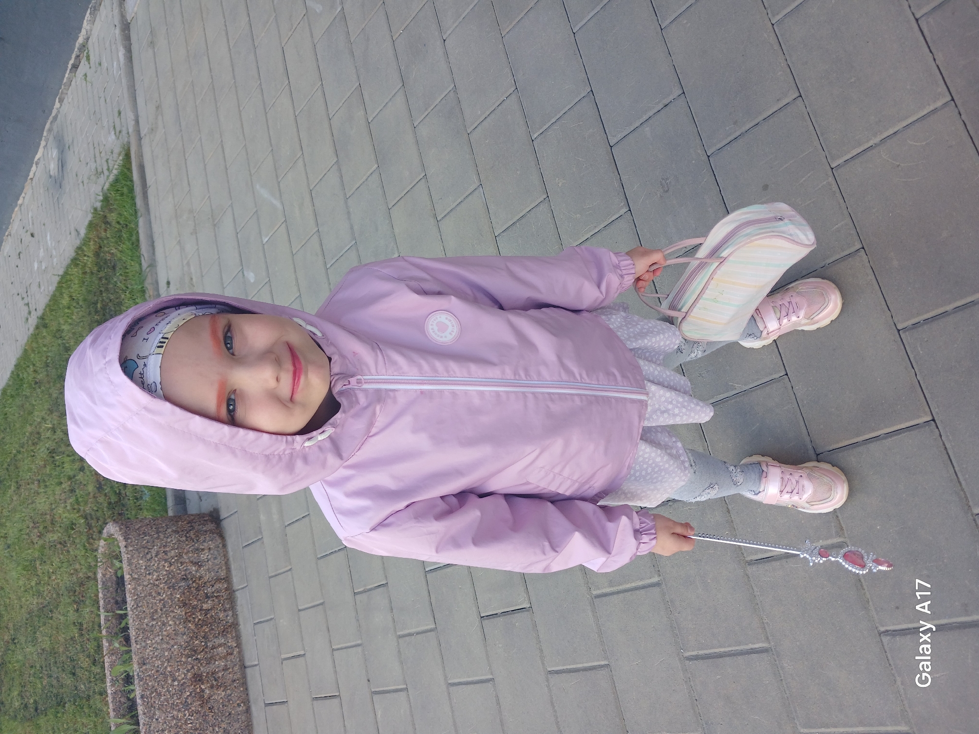 A young girl is standing on a gray brick sidewalk. She wears a pink hooded jacket, a white dress, gray leggings, and pink sneakers. She holds a pastel striped bag in her right hand and a silver wand with pink jewels in her left. A patch of green grass is visible to the left of the sidewalk. Text reading "Galaxy A17" is in the bottom right corner of the image. The image does not contain any indication of the location of Yekaterinburg, Russia.