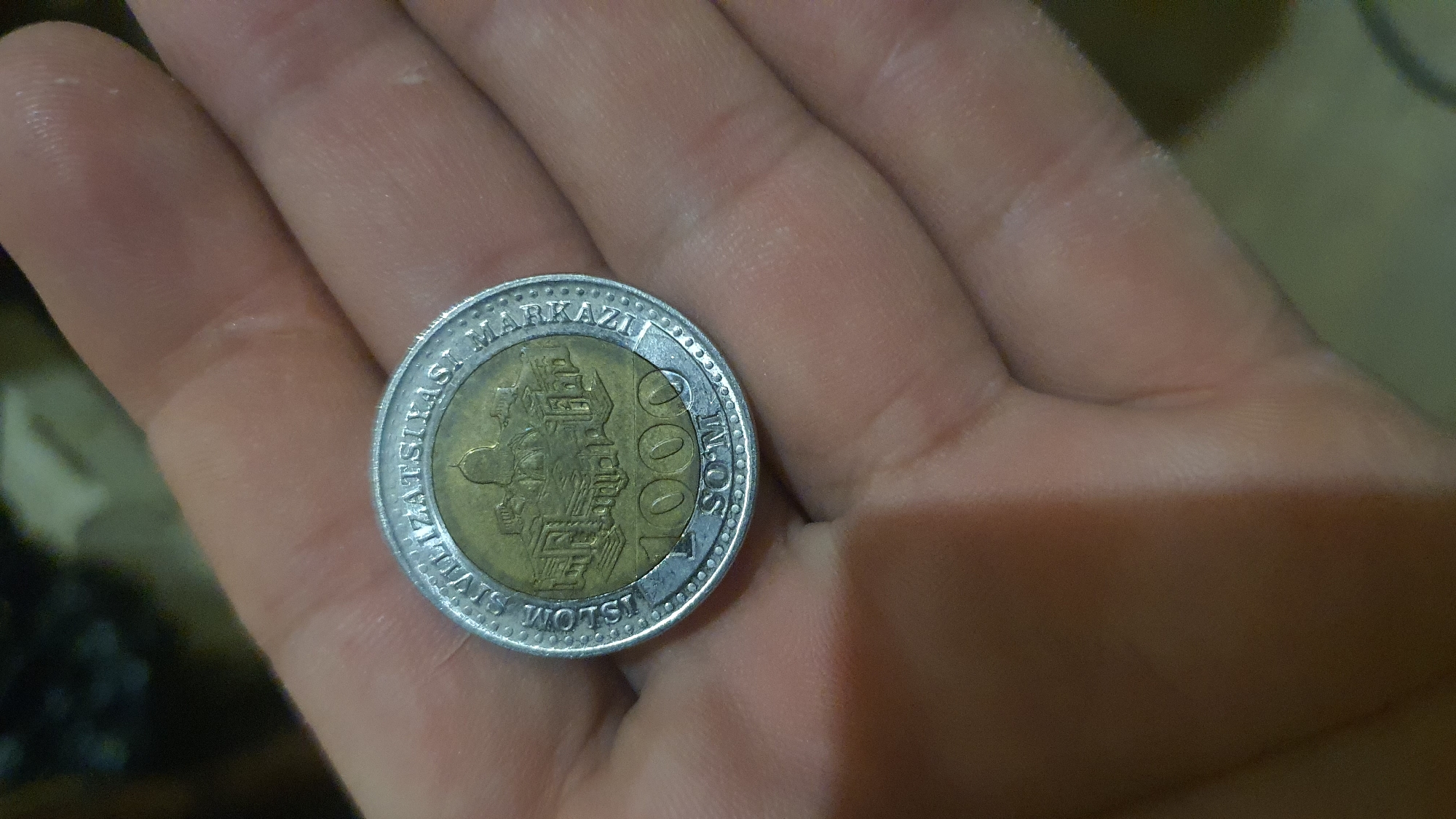A close-up, overhead shot shows a person's hand holding a single coin. The coin has a silver-colored rim and a gold-colored inner circle. The inner circle features an illustration of a building with a dome, surrounded by smaller structures. Text is visible around the rim, reading "ISLOM SIVILIZATSIYASI MARKAZI," and the number "1000" is prominently displayed in the center, followed by "SOM." The coin is resting on the palm of a hand with visible wrinkles and skin texture. The background is blurred and appears to be an indoor setting.