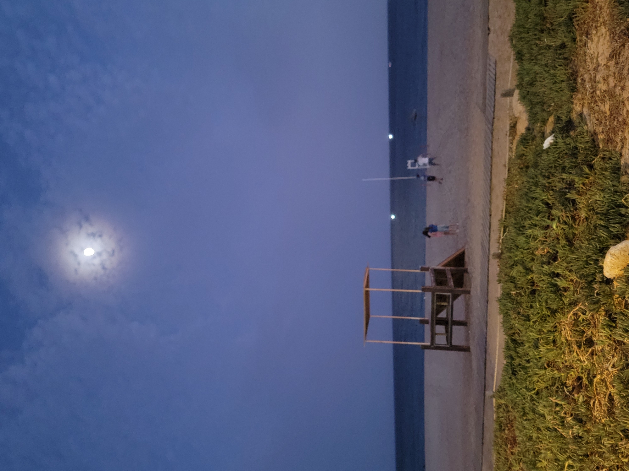 The image depicts a twilight scene at the beach in Quarteira, Portugal. The sky is a deep blue, illuminated by a bright, half-visible moon surrounded by wispy clouds. In the foreground, green vegetation with some dry grasses and a rock is visible. A wooden boardwalk runs horizontally across the middle ground, separating the vegetation from the sandy beach. On the beach, there is a wooden lifeguard tower with a thatched roof. Several people are present on the beach, appearing as small figures in the distance. One person is near the lifeguard tower, and two others are further down the beach. The sea is visible behind the beach, with a dark horizon. Lights are visible in the distance over the water, suggesting distant buildings or boats. The overall impression is a calm evening at the coast.