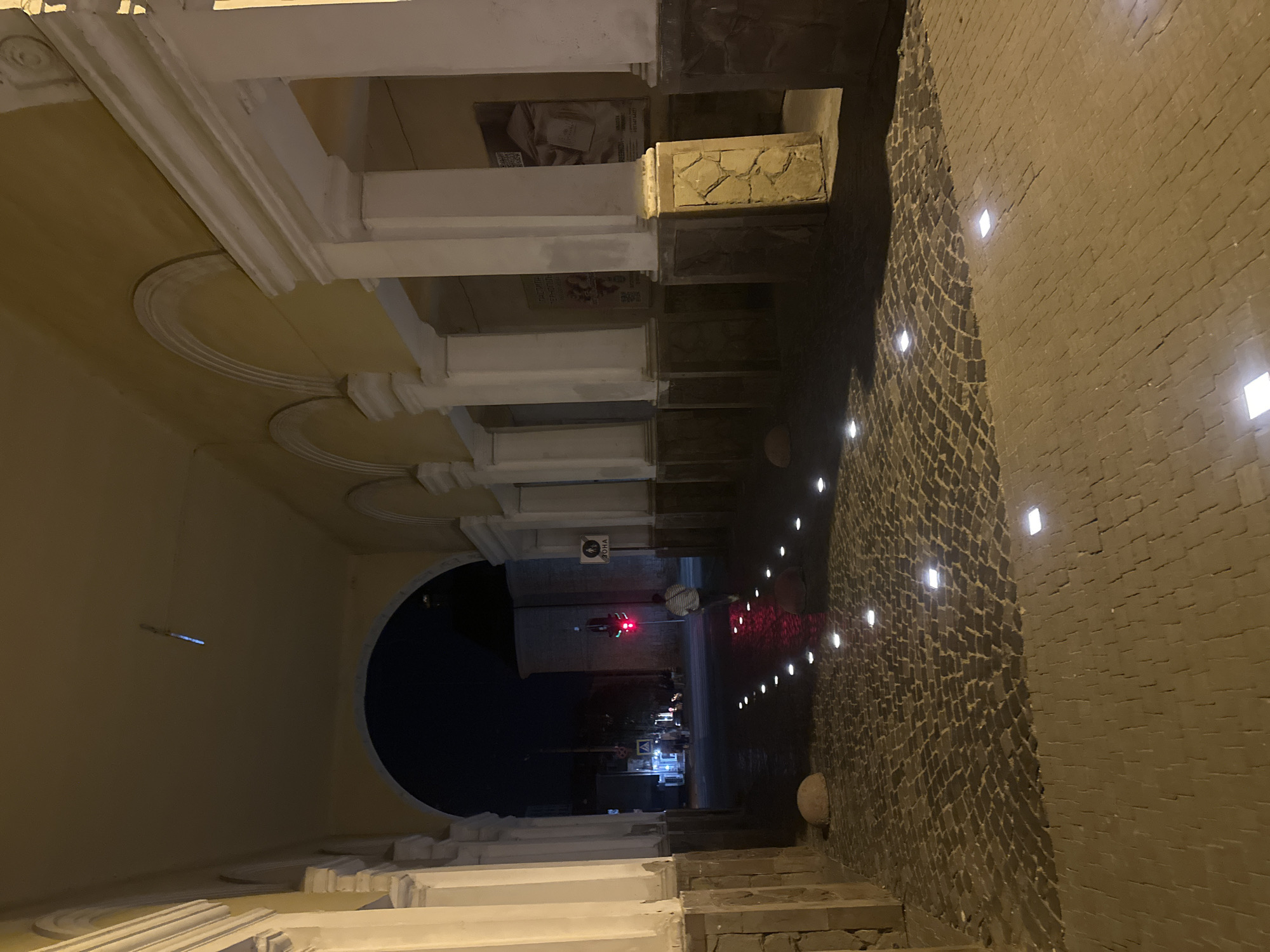 An exterior night shot captures a cobblestone pathway leading through an arched stone and stucco entryway. Embedded square lights illuminate the pathway, creating a dotted line effect. To the right, large white columns with stone bases line the walkway, and advertisements are posted on the wall behind them. In the distance, a traffic light glows red, and a pedestrian walks away from the camera. The pathway is made of small, dark cobblestones and larger, lighter paving stones.
