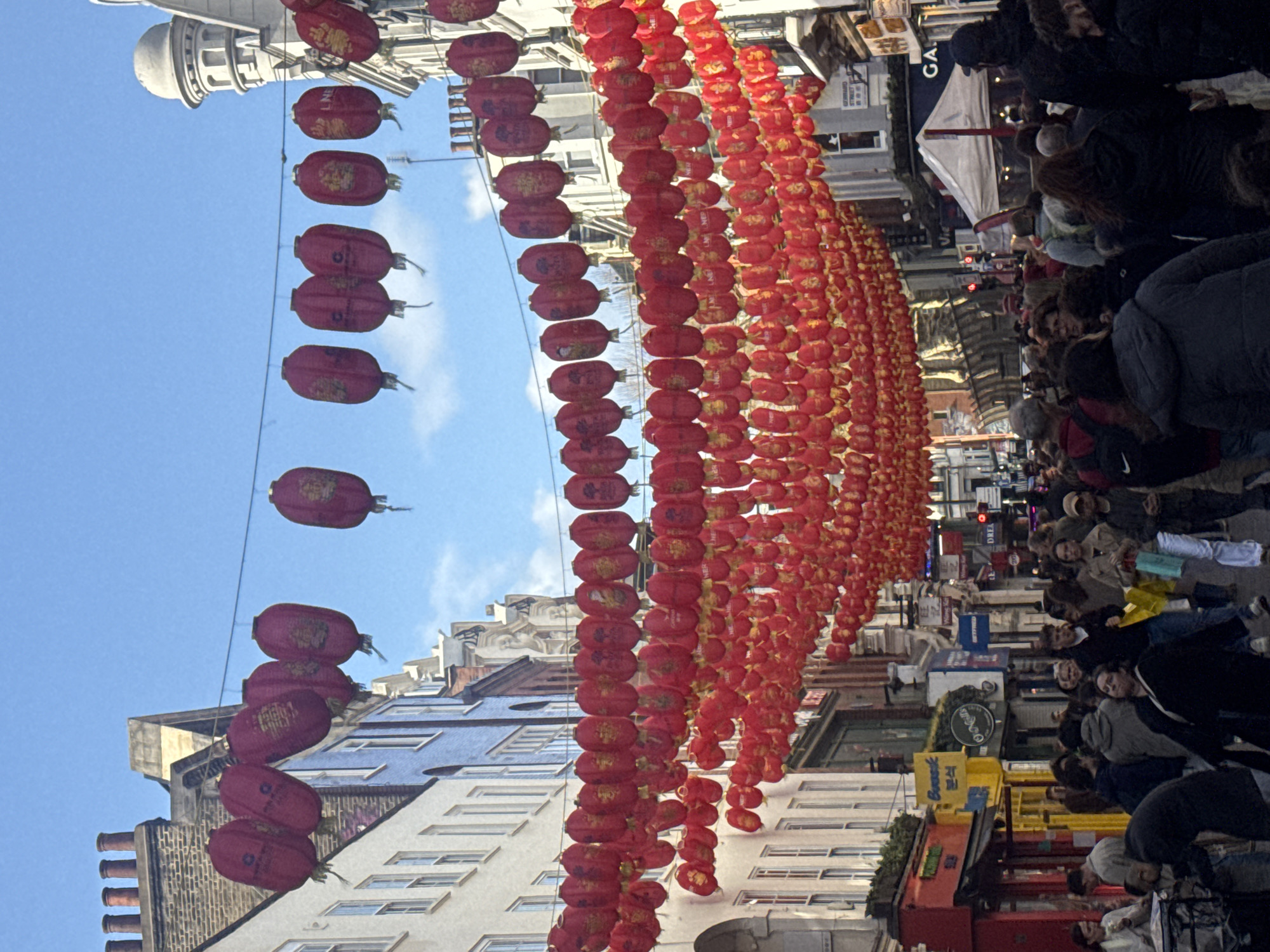 The image depicts a street in London, United Kingdom, likely Chinatown, decorated with numerous strings of red Chinese lanterns. A dense crowd of people is walking along the street, with shops and buildings lining both sides. The sky is visible in the background, appearing clear and blue. The lanterns are the most prominent feature, hanging overhead. The crowd appears to be generally moving forward, possibly sightseeing or attending an event.