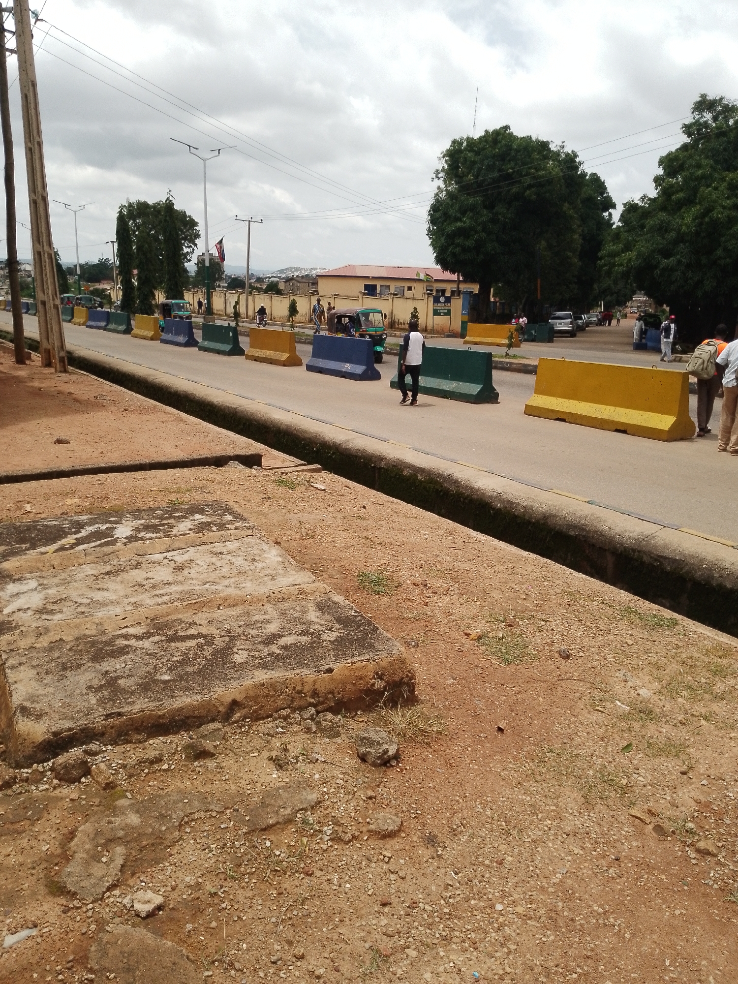 The image depicts a street scene in Jos, Nigeria, featuring a road lined with concrete barriers. The barriers are colored yellow, green, and blue, and are positioned to delineate traffic lanes or a construction zone. Several people are visible walking along the road and on the sidewalk. Some individuals are dressed in casual attire, while others appear to be wearing uniforms. Vehicles, including cars and what appear to be auto-rickshaws or Keke Napeps, are also present on the road. The environment is characterized by a cloudy sky, mature trees, and buildings in the background. The foreground shows a dirt path with some broken concrete structures. The overall impression is a busy urban street with some form of traffic control in place.