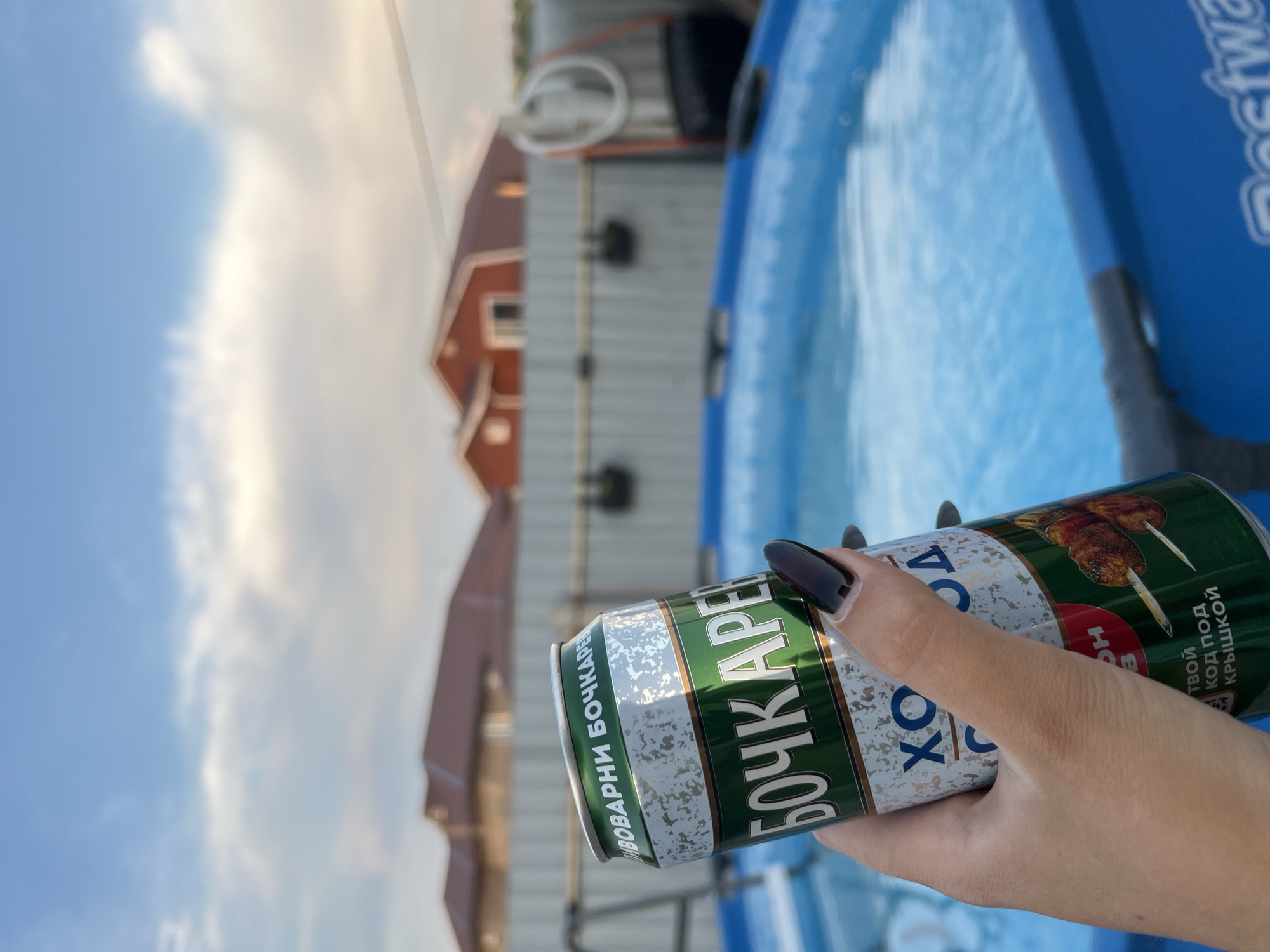 In the city of Solontsy, Russia, a person's hand with dark nail polish holds a green and silver can of beer in front of an outdoor swimming pool. The can has the text "БОЧКАРЕВ" and "ХОЛОД" on it, and features an image of meat skewers. The background shows a residential area with houses and a fence, under a cloudy sky. The pool appears to be filled with water and a portion of the "Bestway" logo is visible on its edge.