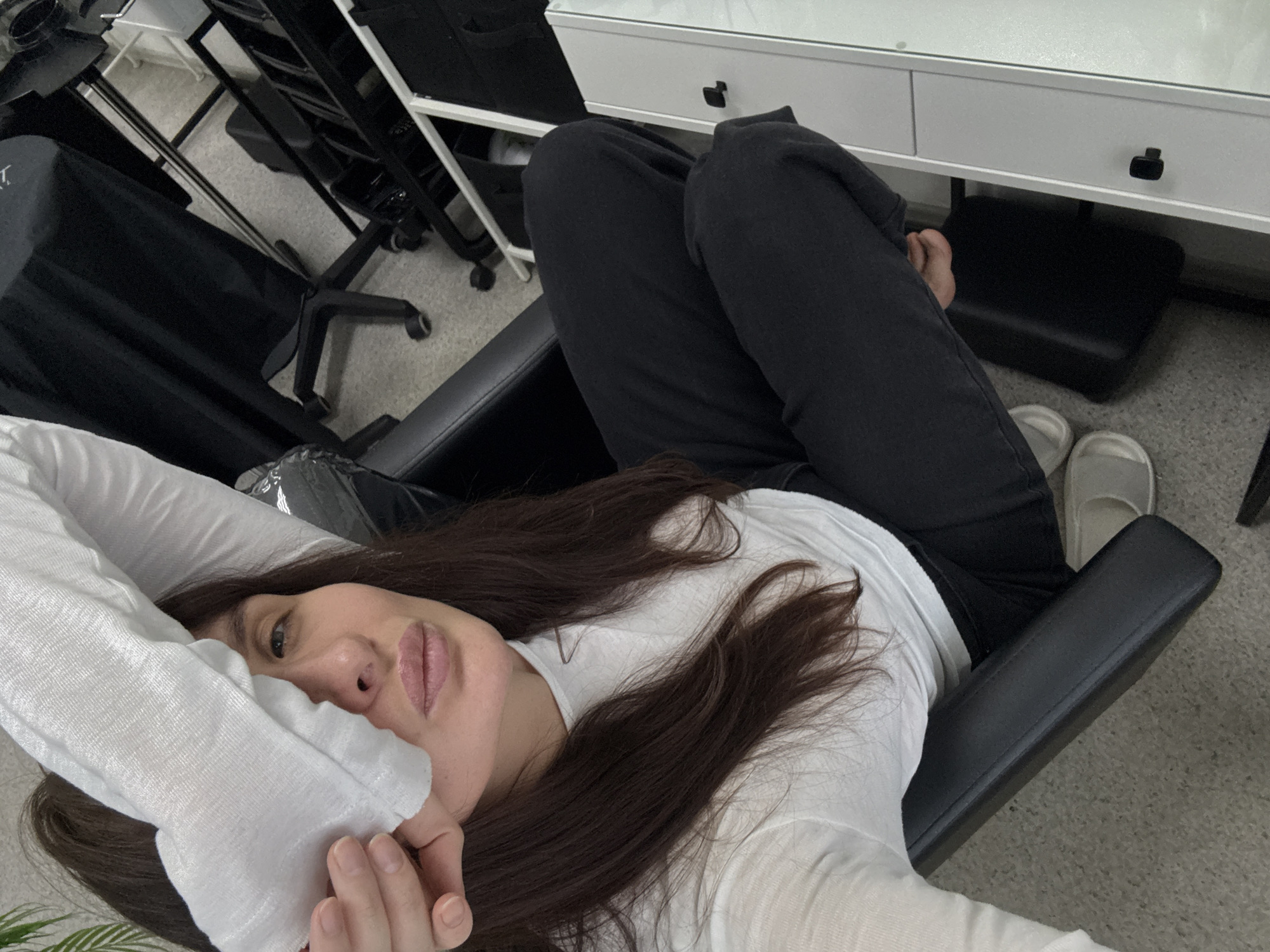 A person lies in a black chair, wearing a white shirt and black pants, with grey slippers on the floor nearby. The person has long brown hair and is partially obscuring their face with their hand and shirt. A white vanity with black drawer pulls is visible in the background, along with a rolling black rack. The location is Chaikovsky, Russia.