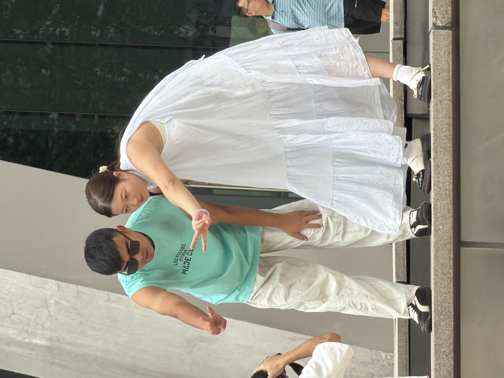 In Yuzhong District, China, a man wearing sunglasses and a turquoise sleeveless shirt stands next to a woman in a light blue tiered dress. The man is leaning forward, looking down and gesturing with his left hand. The woman is also looking down and holding up her right hand in a peace sign. Both are on a raised stone platform. In the background, a man in a blue striped shirt and shorts is visible. The environment appears to be outdoors with a modern architectural style, suggested by the smooth concrete walls and reflective dark green panels.