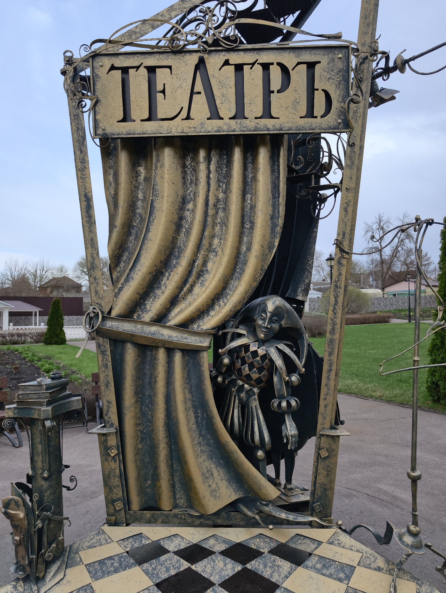 The image depicts a metallic sculpture in Balashov, Russia. The sculpture is an artistic representation of a theater entrance, with a sign reading "ТЕАТРЪ" (Theater in pre-reform Russian orthography) above a simulated curtain. A harlequin figure is partially visible behind the curtain. The sculpture is located in an outdoor park setting with grass, trees, pathways, and buildings visible in the background. Other metallic objects, including a representation of books and a coat hanger, are attached to the main sculpture. The base of the structure features a checkered pattern. The overall style of the sculpture is ornamental, using a mix of gold and black coloring.
