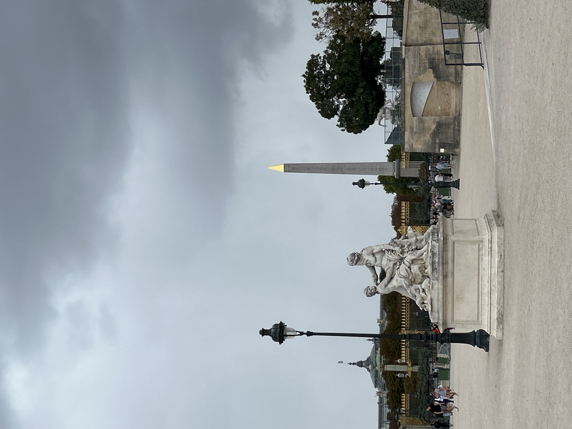 This is a vertical, eye-level, medium-shot photograph of the Place de la Concorde in Paris, France, on an overcast day. In the foreground, a white stone statue of two figures is on a pedestal. To the left of the statue is a black lamppost. In the middle ground, a tall, gray obelisk with a gold cap stands prominently. Beyond the obelisk, there is a large, dark green tree. To the left of the obelisk, a golden-capped dome building is visible, behind a golden fence. The ground in the foreground is a light beige gravel. Several people are walking in the middle ground and to the left of the statue.