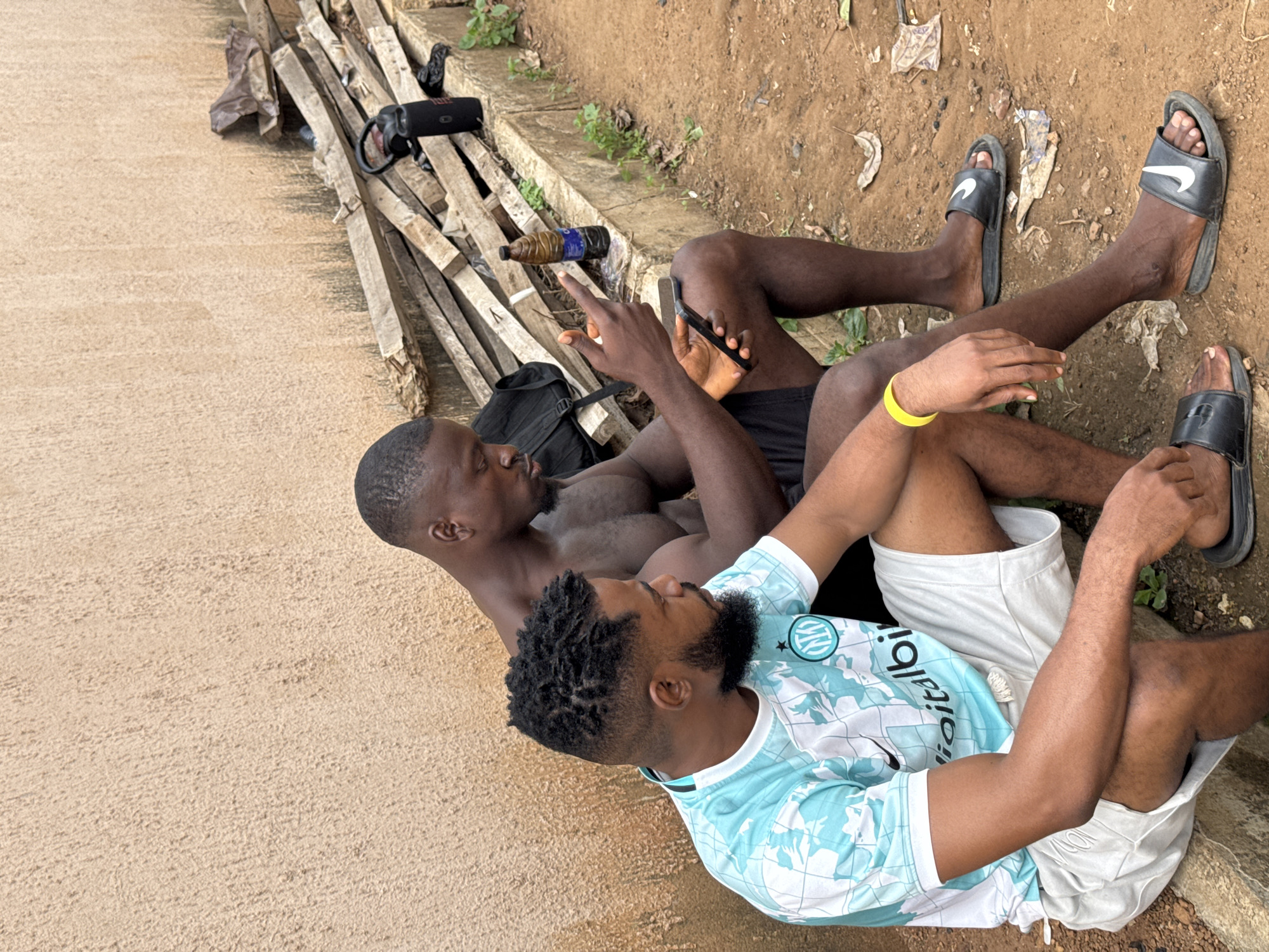 Two men are seated on a concrete ledge outdoors. The man on the left wears a light blue and white patterned t-shirt and white shorts, with a yellow wristband on his left arm. He has dark, curly hair and a beard. The man on the right is shirtless and wears black shorts. He has short dark hair and is looking down at his phone, which he holds in his right hand. His left hand is raised, with his index finger extended. Beside him, on the ledge, are a black backpack, a bottle of liquid, and a black water bottle. The background consists of a textured, light brown wall. The ground in front of the ledge is dirt. In the upper right of the image, a pile of wooden planks is visible. This scene is located in Akwanga, Nigeria.