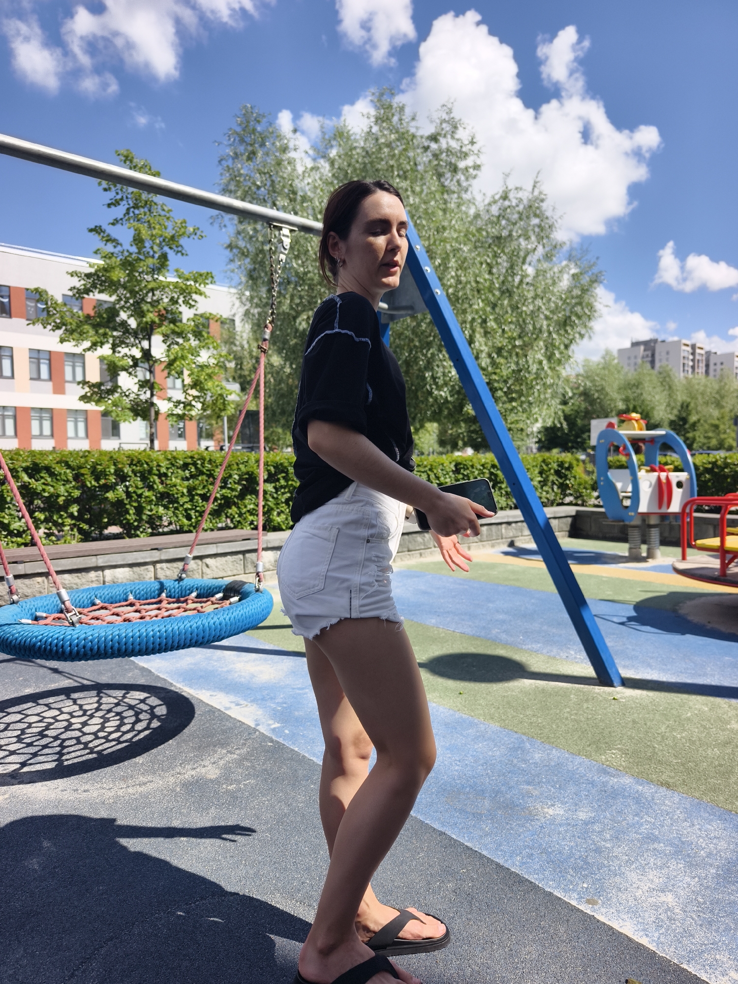 A young woman stands on a playground, holding a phone in her right hand and looking over her left shoulder. She is wearing a black t-shirt with white trim and white denim shorts. The playground features a large blue nest swing, a blue metal swing frame, and a multi-colored play structure in the background. The ground is a rubberized surface with blue, green, and grey sections. Behind the playground, there are buildings and trees, suggesting an urban setting. The sky is blue with scattered white clouds. The scene is illuminated by bright sunlight, casting shadows on the ground. The location is identified as the city of Tyumen, Russia.