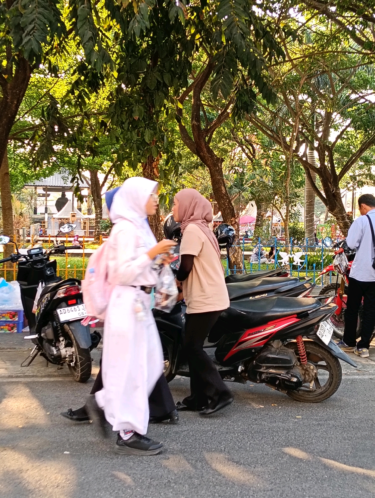 Two young women, both wearing hijabs, are walking on a street in Karanganyar, Indonesia, next to several parked motorcycles. The woman on the left is wearing a long white robe, a blue hijab, and a pink backpack. The woman on the right is wearing a beige shirt and a pink hijab. They appear to be interacting, with the woman on the left holding a clear plastic bag. Behind them, a man in a light blue shirt and dark pants is standing next to a red motorcycle. The background shows trees, a fence with decorative elements, and some buildings. The scene is illuminated by sunlight, casting shadows on the asphalt street.