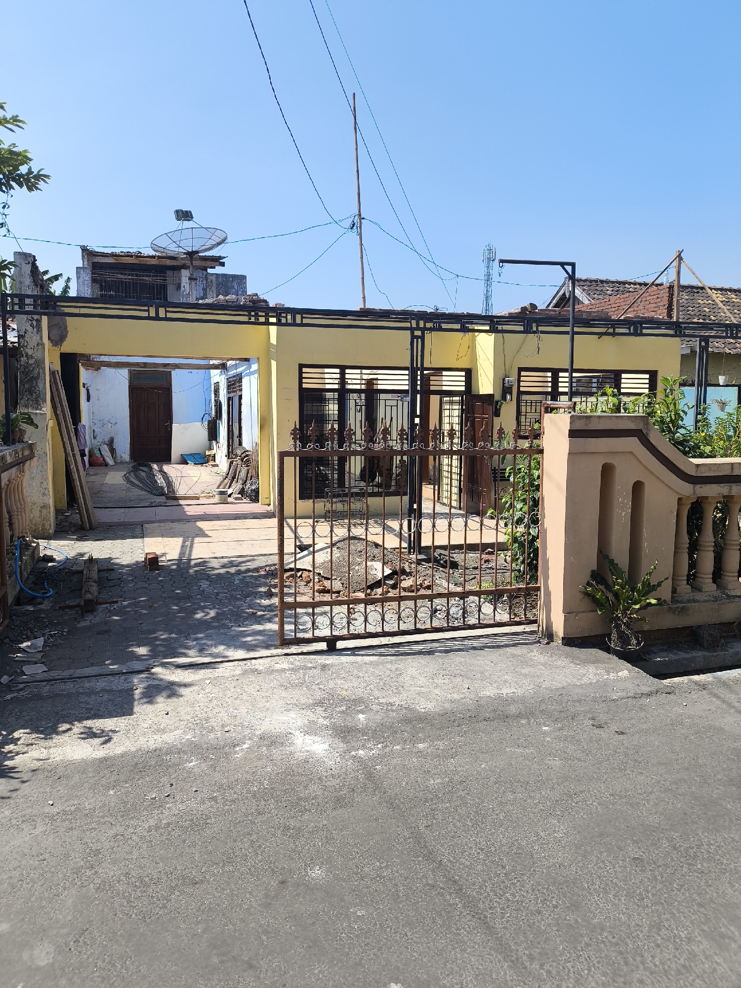 A residential house with a yellow facade is visible from the front, under a clear blue sky. A rusty metal gate with circular ornaments stands in front of the property. The gate is slightly ajar, revealing a partially paved courtyard with debris, including bricks and construction materials. Beyond the gate, the house features a covered porch area leading to an interior with a wooden door. The house has several windows with horizontal blinds. A satellite dish is mounted on a rooftop structure behind the house, along with a telecommunications tower in the distance. The street in front of the property is asphalted and shows signs of wear. The environment appears to be a sunny day in a residential area, possibly undergoing some renovation or construction.