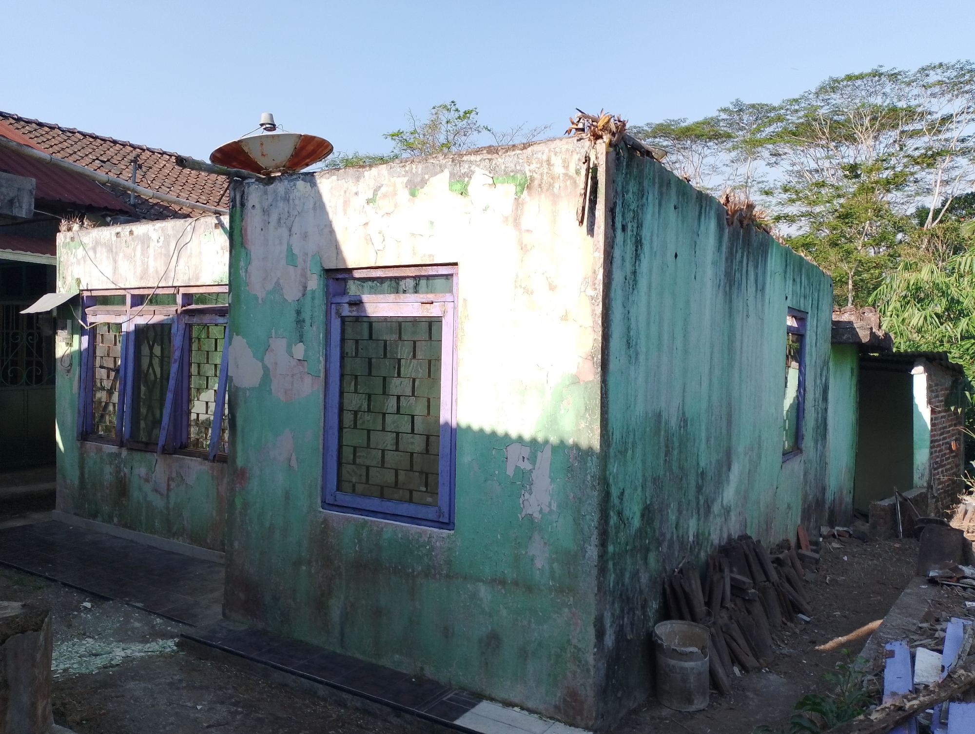 The image shows an exterior view of a partially damaged, abandoned building in the city of Payaman, Indonesia. The building has a dilapidated appearance, with peeling green and white paint on its walls. It features several windows with purple frames, some of which appear to be boarded up or have bars. A satellite dish is mounted on a concrete pillar to the left of the building. In the foreground, there is debris scattered on the ground, including wood and what appears to be roofing tiles. Piles of wood are stacked against the wall of the building. The environment is outdoors, with a clear blue sky above and lush green trees and vegetation in the background. The scene suggests a rural or semi-rural setting. There are no people visible in the image.