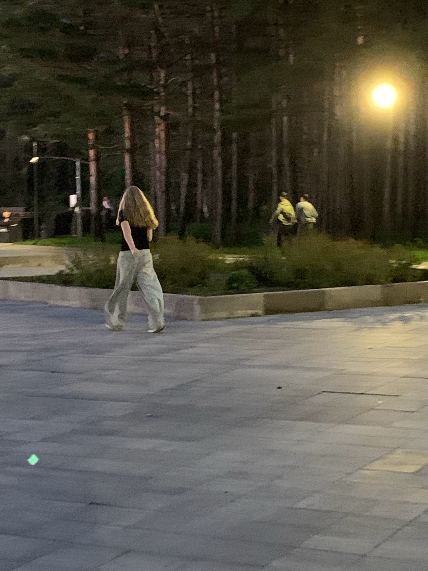 The image shows an outdoor scene at dusk, likely in a park or a public space with paved ground and trees. In the foreground, a person with long blonde hair, wearing a dark t-shirt and light-colored baggy pants, is walking away from the camera. In the background, two individuals wearing light-colored jackets are walking in the same direction, further into a wooded area. A bright light source, possibly a streetlamp or a moon, is visible in the upper right portion of the frame, illuminating the scene. The environment appears to be a moderately vegetated area with tall trees. The presence of paved walkways suggests a developed public area. Without further context or landmarks, it is impossible to definitively pinpoint the location to Prokopyevsk, Russia, but the scene depicts a common urban or suburban outdoor environment.