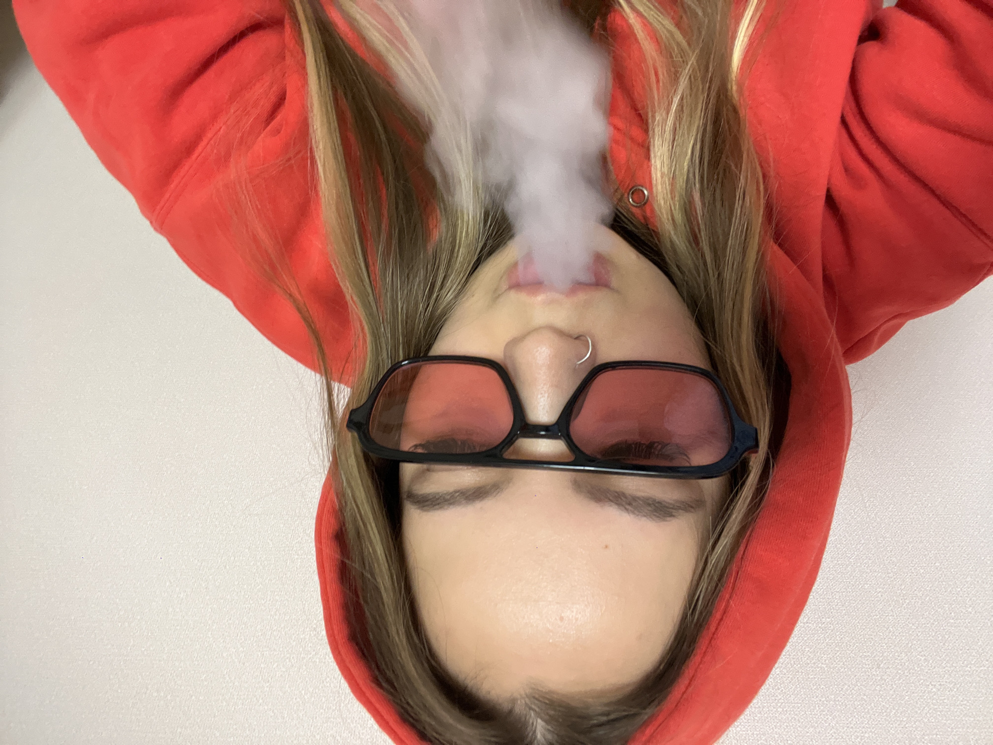 A young person with blonde hair and wearing a red hoodie is looking up at the camera. They are wearing black-framed sunglasses with tinted lenses, and there is a nose ring in their left nostril. The person is exhaling a large puff of white smoke. The background is a plain white surface. No evidence in the image to suggest the location is Krasnodar, Russia.