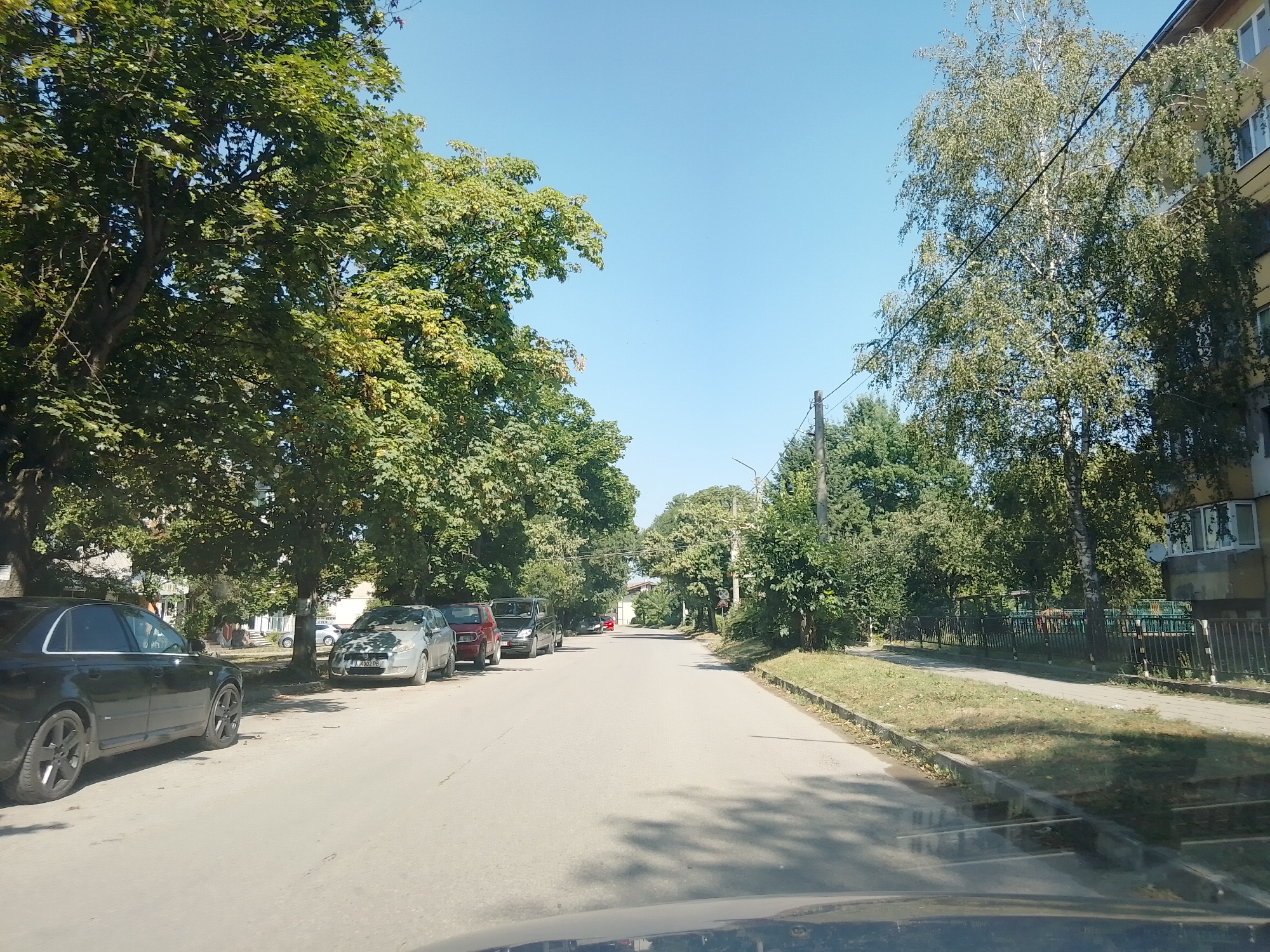 The image depicts a sunny day scene on a residential street in Ihtiman, Bulgaria. The road is paved and appears to be two-lane, with parked cars lining the right side of the street. On the left side, a black sedan is prominently parked, followed by other vehicles including a silver hatchback, a red SUV, and a dark van. Lush green trees with dense foliage overhang the left side of the road, casting shadows. To the right of the road, a sidewalk is visible, separated by a grassy verge. Beyond the sidewalk, a black metal fence runs along the property line of a building that appears to be an apartment complex, characterized by a yellow facade and several windows. Another tall birch tree with light bark stands near the apartment building. The sky is a clear, bright blue with no clouds. The overall impression is a typical, tranquil urban residential environment.