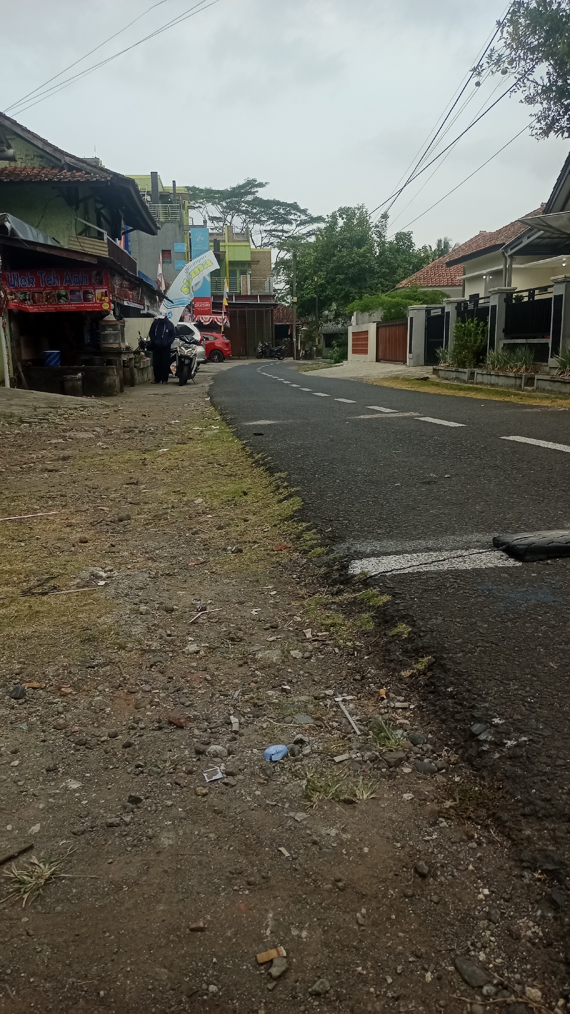 The image depicts a scene on a paved road in Pakemitan, Indonesia. The road has white dashed lines indicating lanes. On the left side of the road, there is a roadside structure with a red sign that reads "Ulek Teh Aah." A person in a dark jacket and pants is standing next to a motorcycle. Behind them, a red car is parked, and further down the road, another motorcycle is visible. The area to the left of the road is unpaved and rocky, with some sparse grass and debris, including a blue plastic bottle and cigarette butts. On the right side of the road, there are houses with tiled roofs and fences. The sky is overcast.