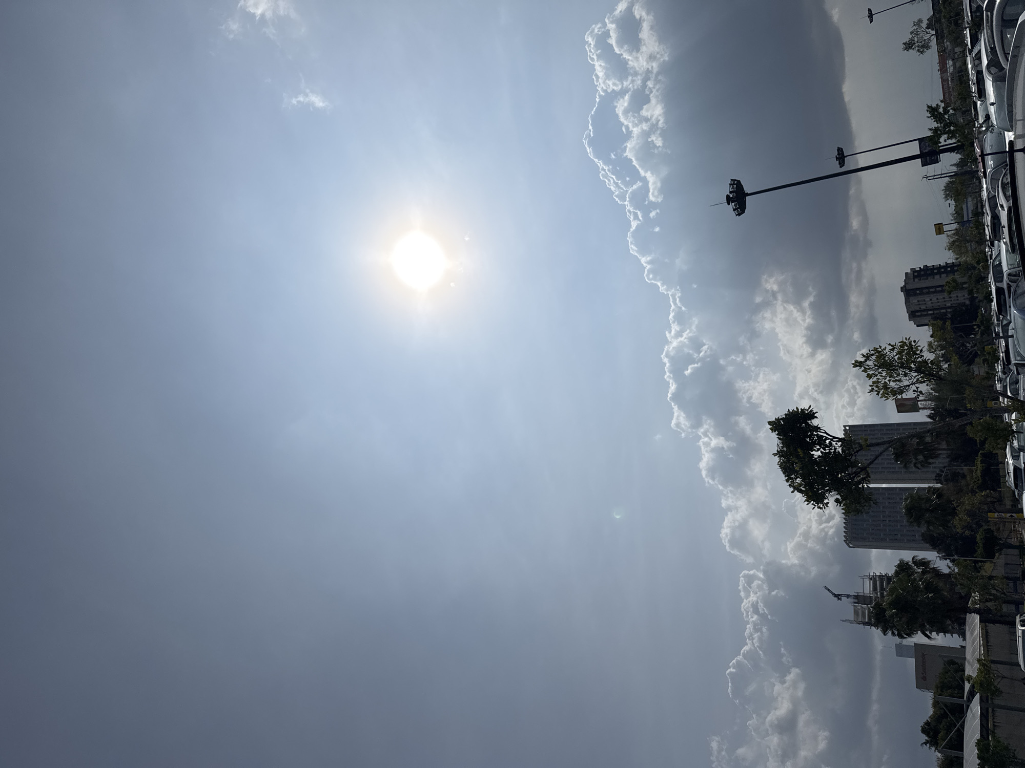 The image depicts a daytime view of the sky in Puebla, Mexico, characterized by the sun shining through a mix of clear skies and cumulus clouds. The city skyline is visible on the right side of the image, featuring buildings, trees, lampposts, and what appears to be a parking lot full of cars. The sun is positioned centrally in the frame, while the clouds are predominantly on the right.