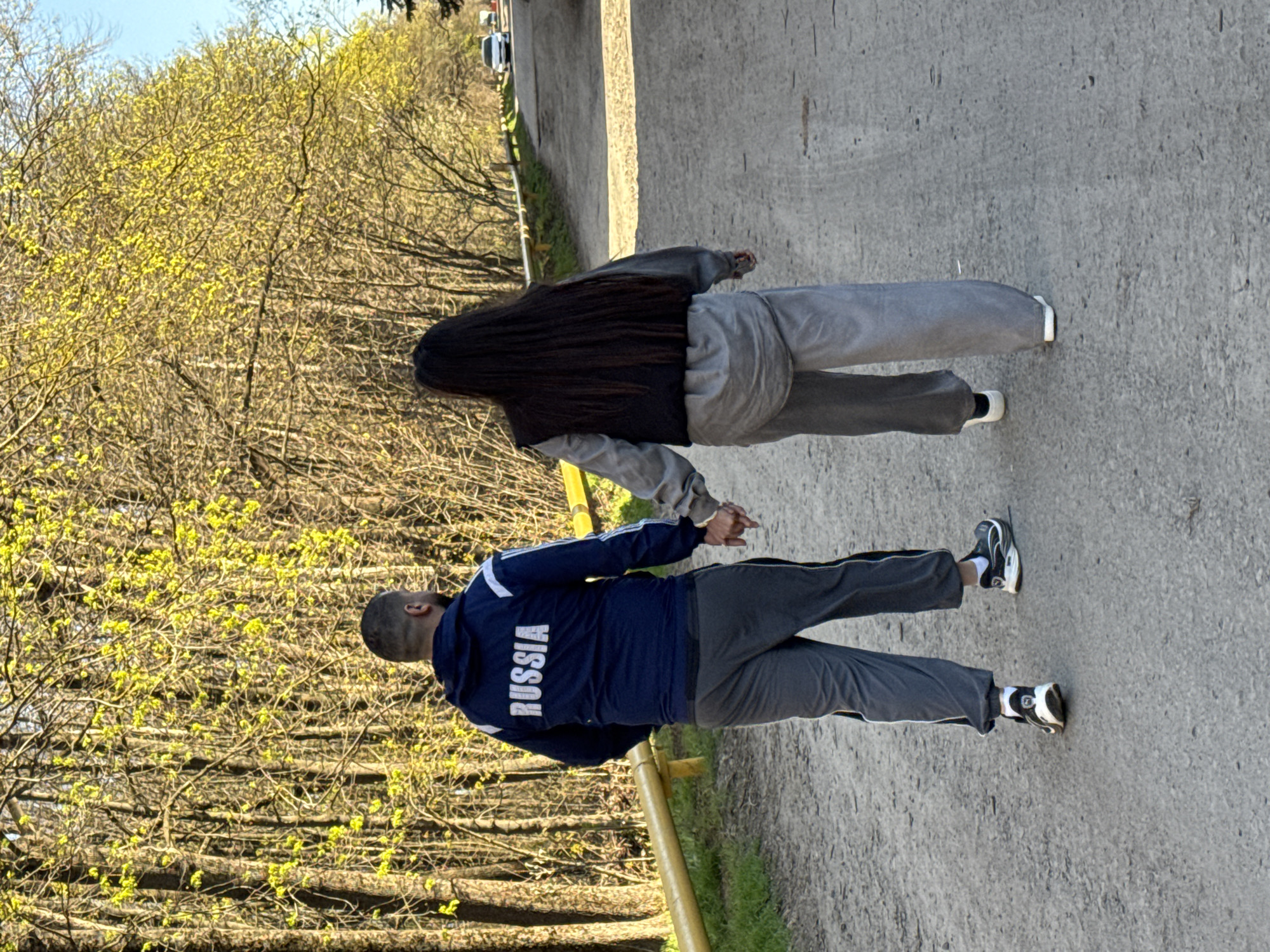 Two individuals are walking along a concrete path. A man wearing a dark blue jacket with "RUSSIA" printed on the back and dark grey pants is holding the hand of a person with long, dark hair wearing a grey sweatshirt and grey pants. Trees with early spring foliage line the path. The location is likely Rostov-on-Don, Russia.