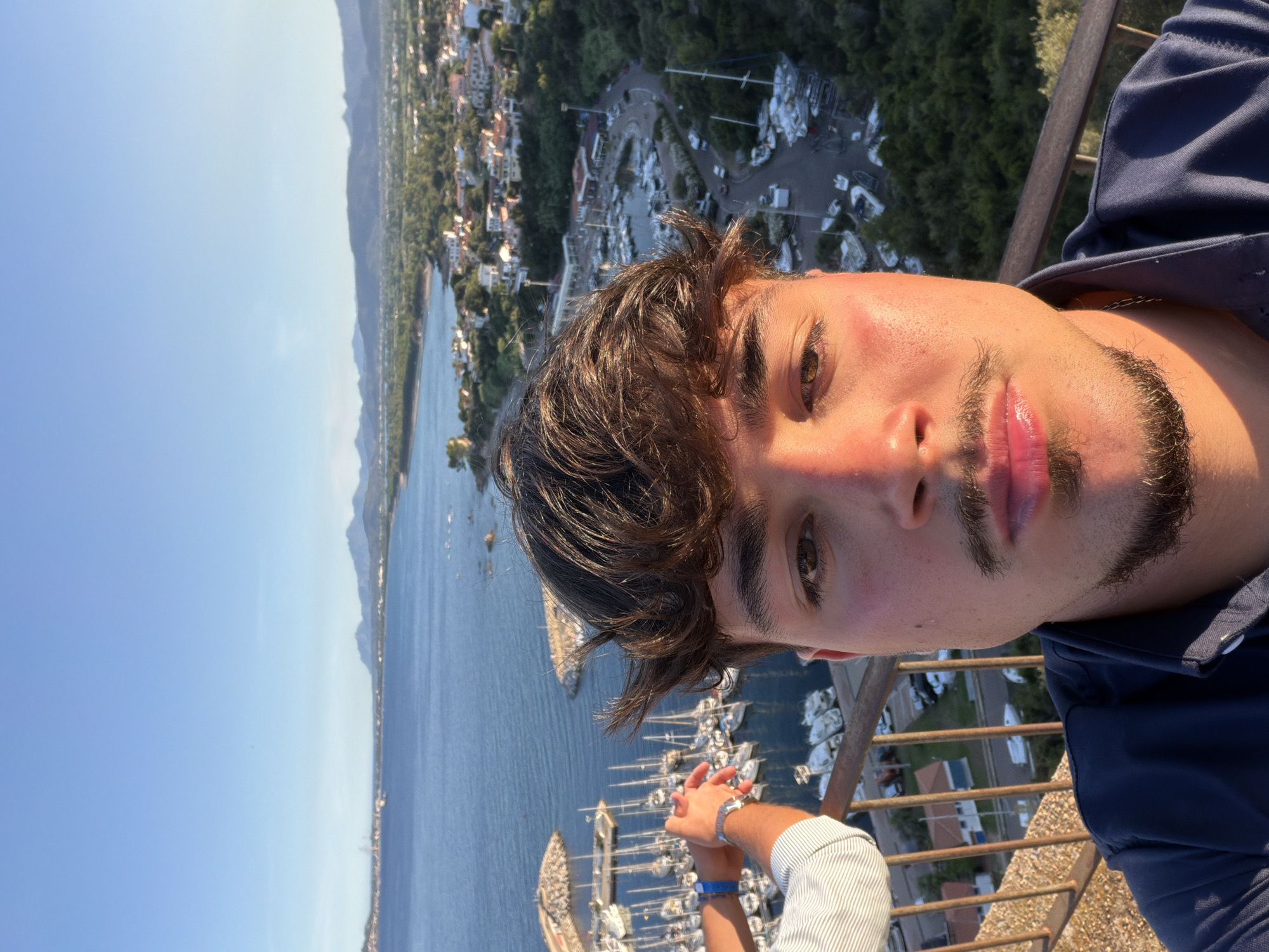 The image is a selfie taken by a young man with brown hair and a mustache, standing on a balcony overlooking a harbor in Santa Maria Navarrese, Italy. The man is wearing a dark blue shirt. To his left, another person's arms are visible, one wearing a striped shirt and a watch, the other with a blue bracelet.
The background reveals a picturesque scene of a bay filled with numerous white sailboats docked. Beyond the harbor, the coastline is dotted with buildings and lush green vegetation, leading to rolling hills and distant mountains under a clear blue sky. The sun casts a warm glow on the man's face, indicating it is likely late afternoon or early evening. The perspective suggests the photographer is positioned at a height, offering a panoramic view of the coastal town and its marina.